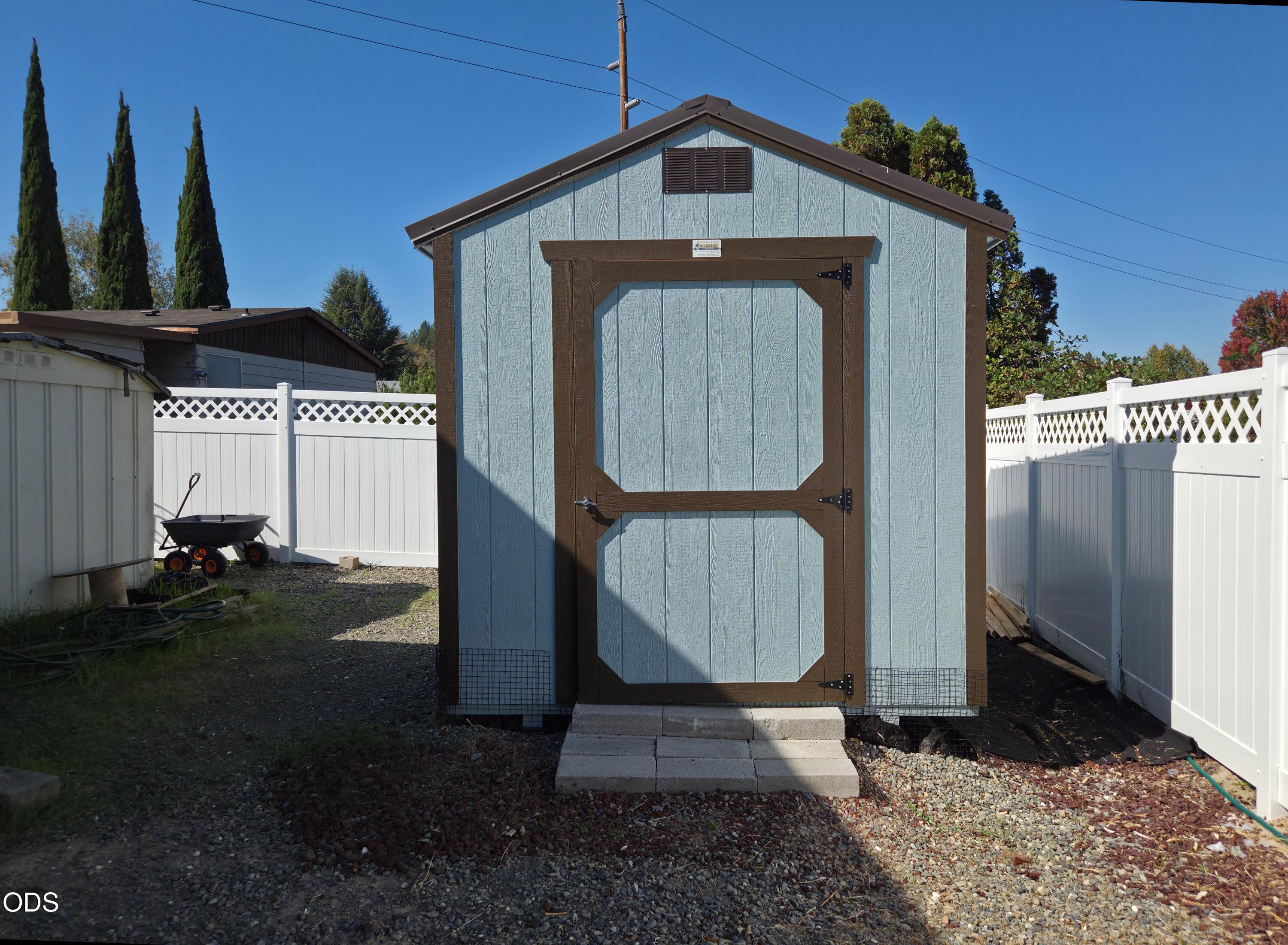 Blue garden shed with brown trim, set between white fences in a gravel yard.