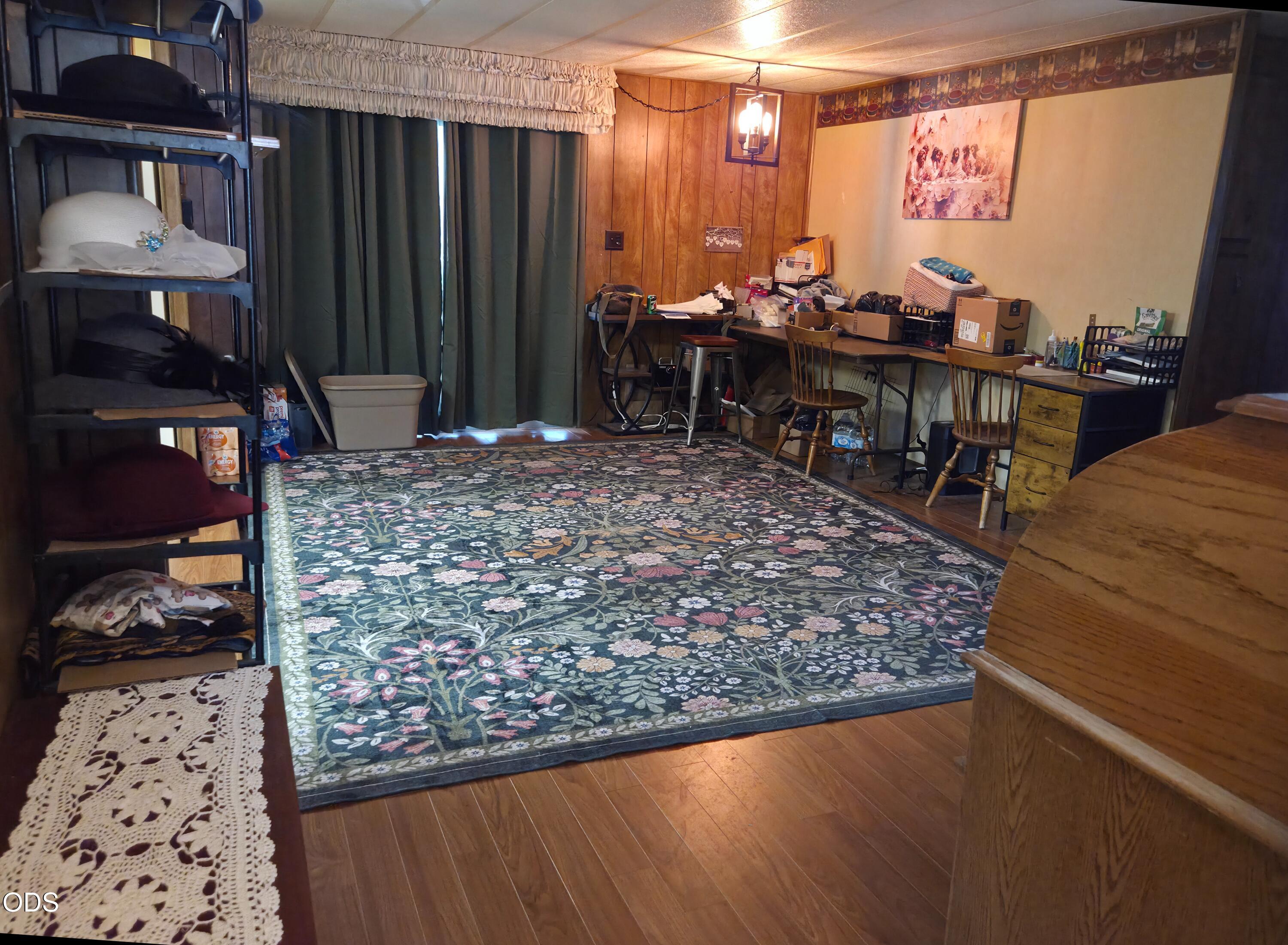 Cluttered home office with a large floral rug, long worktables, and wood-paneled walls.