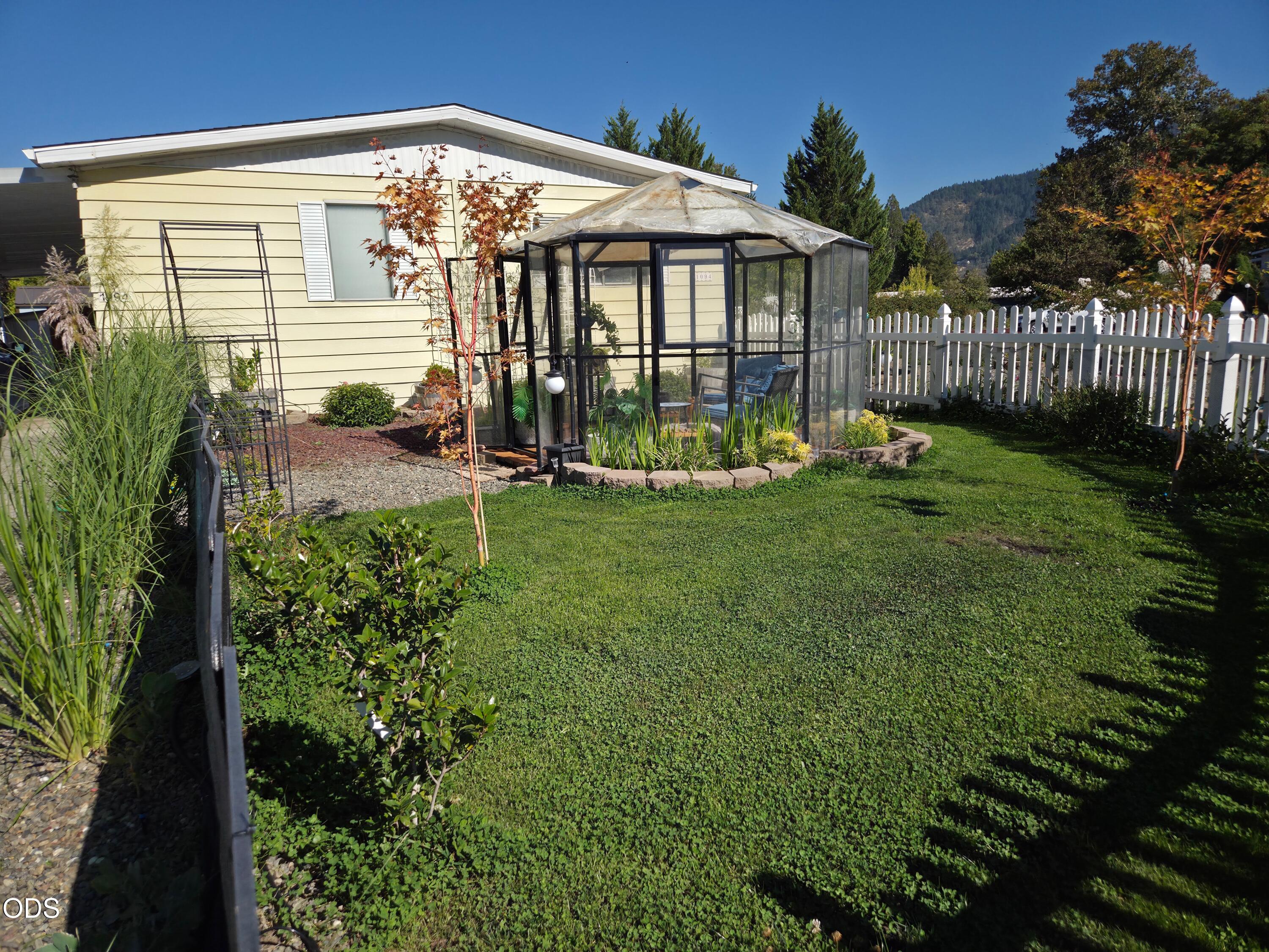 Backyard with a glass greenhouse beside a pale yellow home and white picket fence.