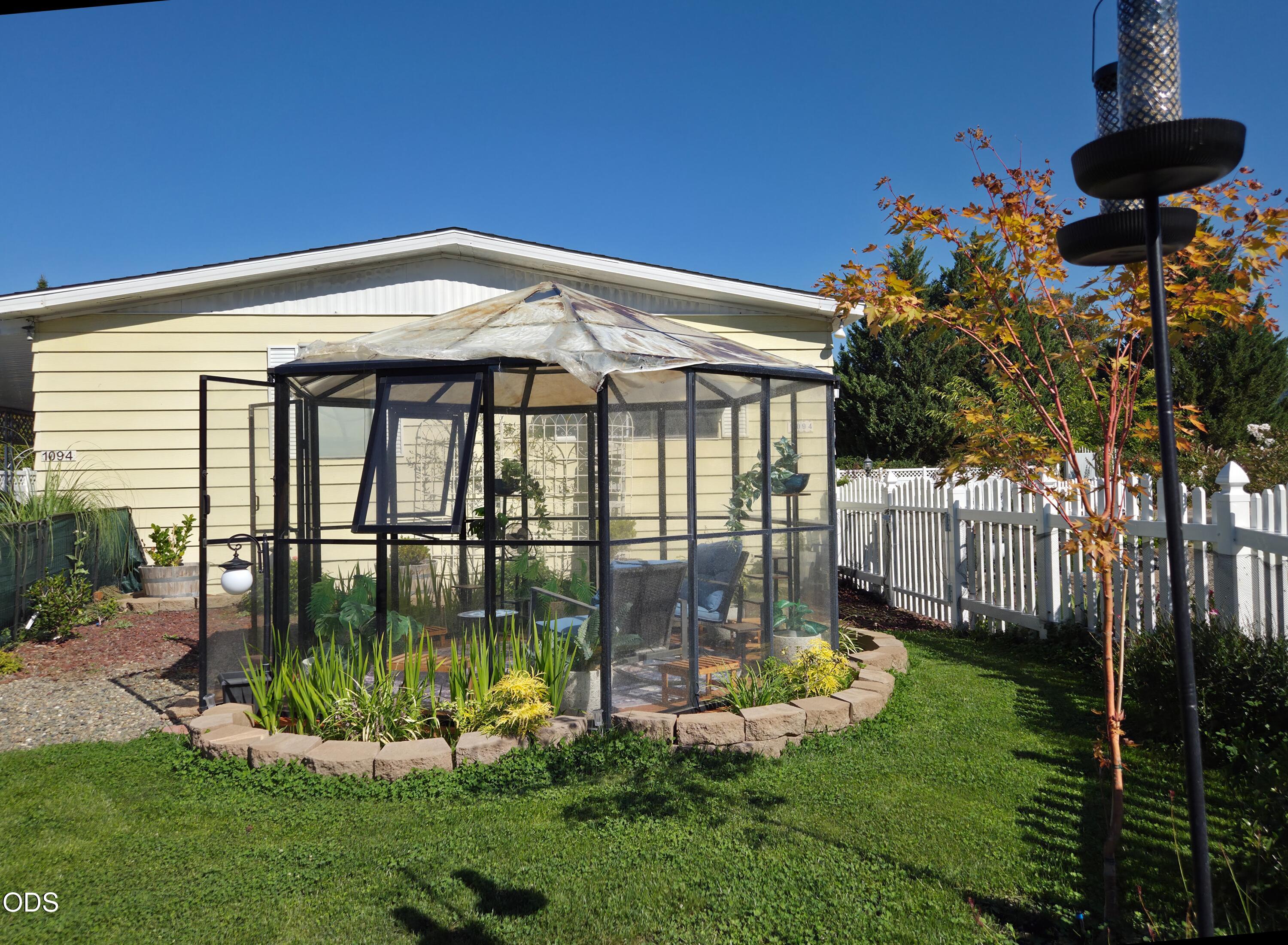 Backyard greenhouse beside a pale yellow home, with a white picket fence and autumn foliage.