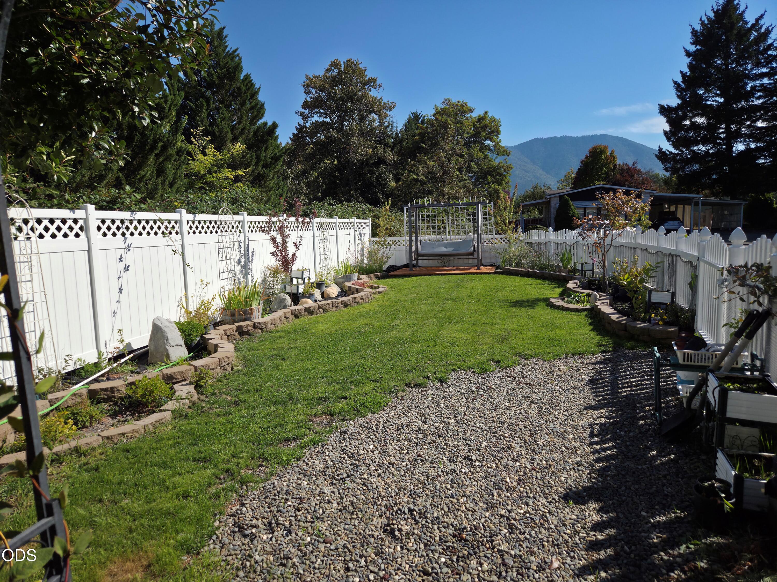 Fenced backyard with a lawn, gravel path, garden beds, and a pergola swing beneath mountain views.