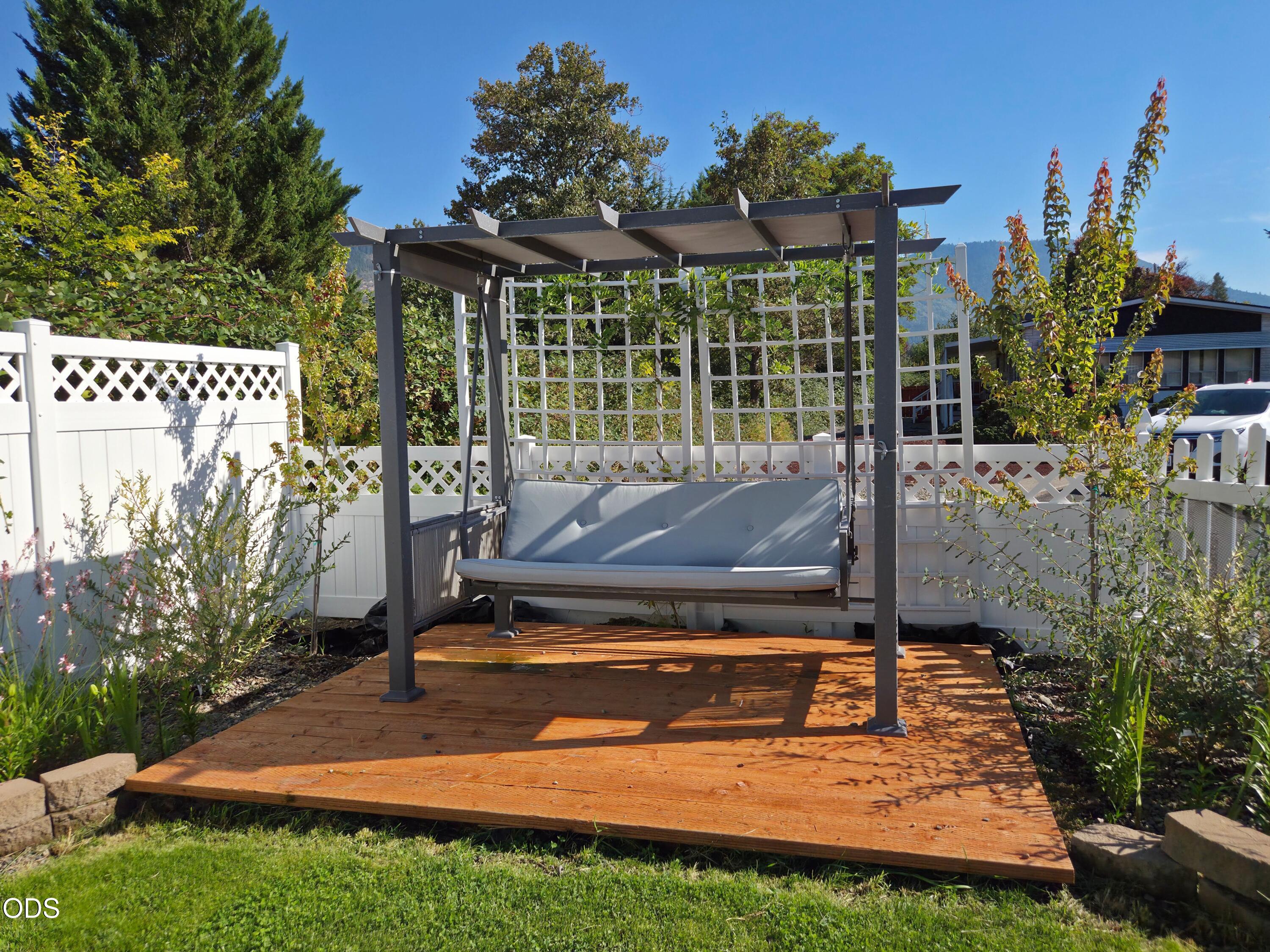 Wood deck with a cushioned swing beneath a pergola, surrounded by garden plantings and white fencing.