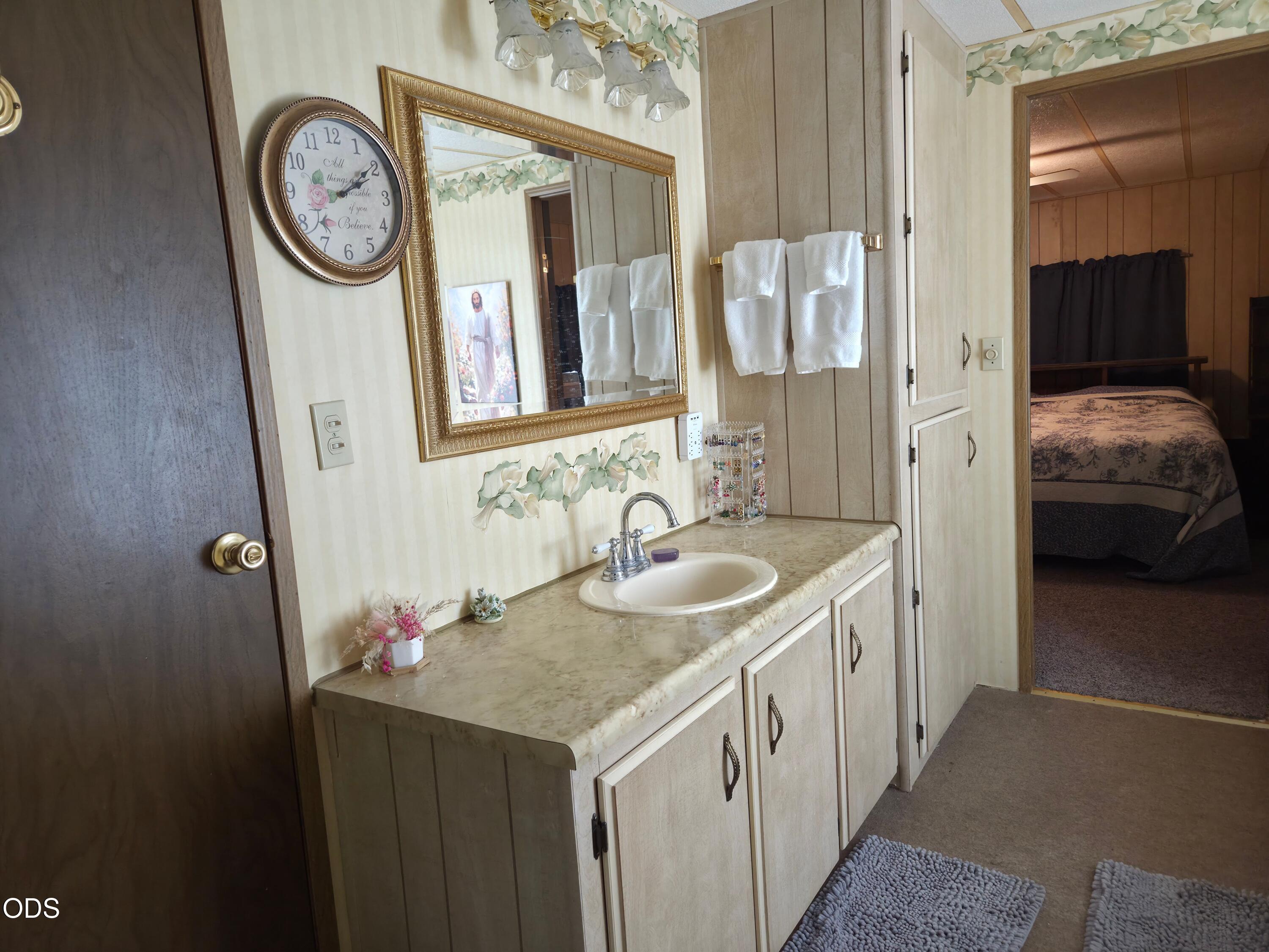 Bathroom vanity with a marble-look counter and gold-framed mirror, with a bedroom visible through the doorway.