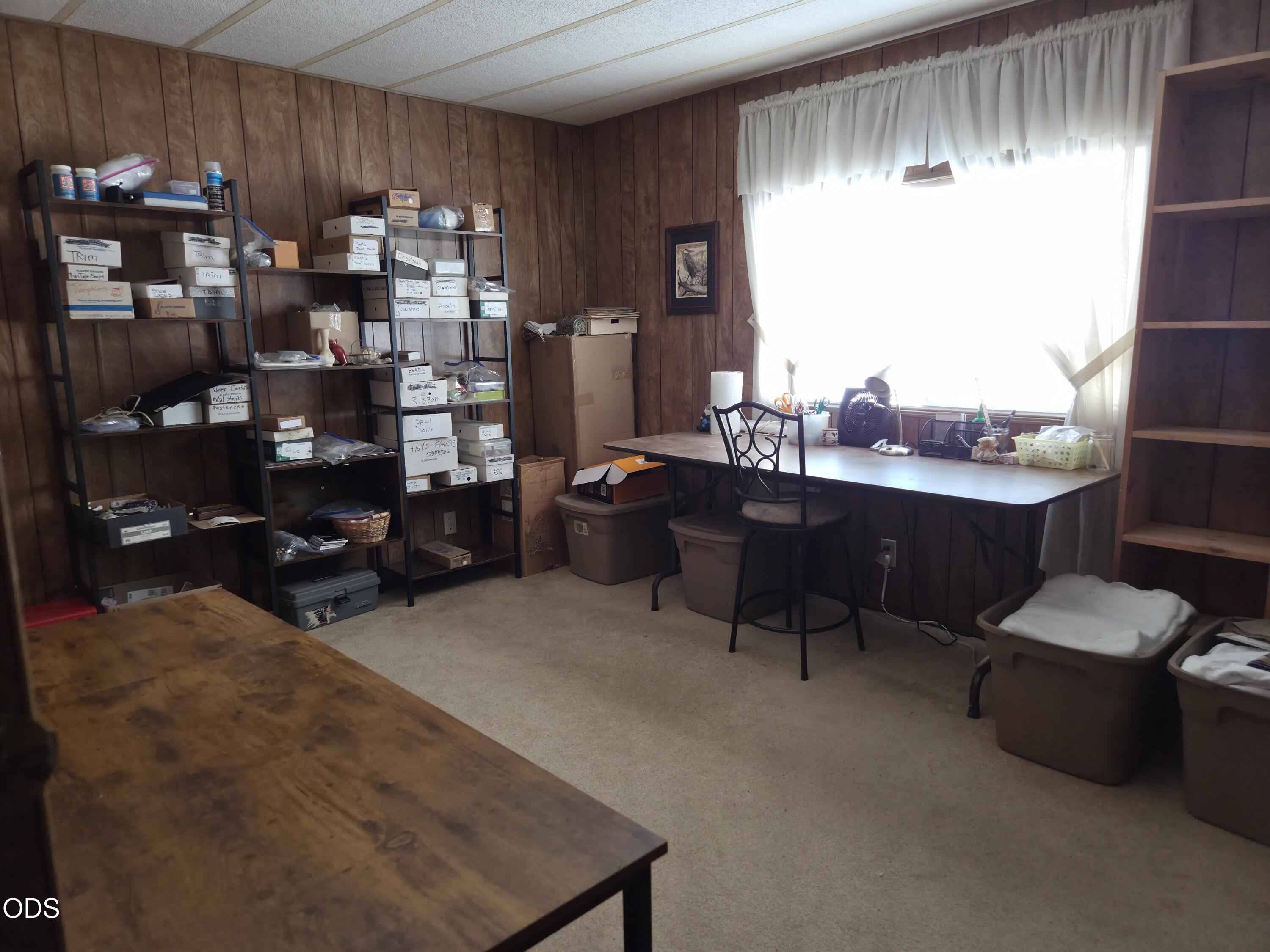 Cluttered office and storage room with wood-paneled walls, shelving, and a desk by a bright window.