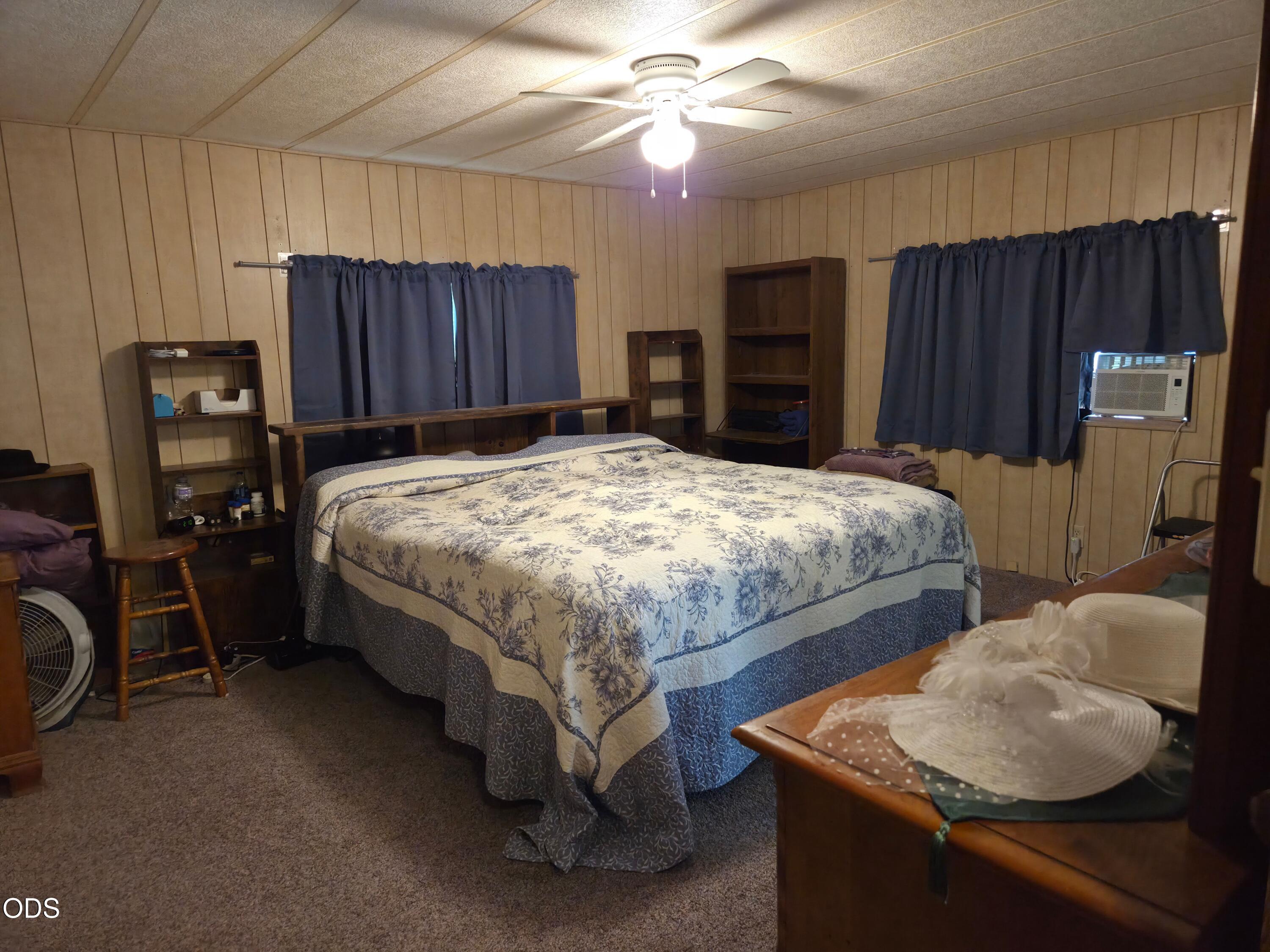 Bedroom with a floral quilted bed, wood-paneled walls, dark curtains, and a window air conditioner.