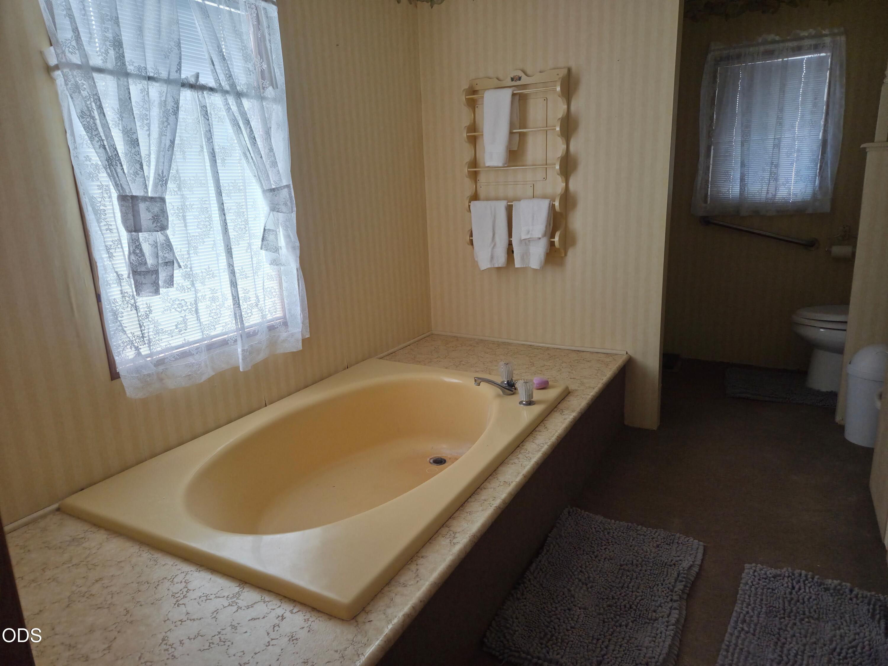 Bathroom with a cream soaking tub beneath a lace-curtained window and a separate toilet area.
