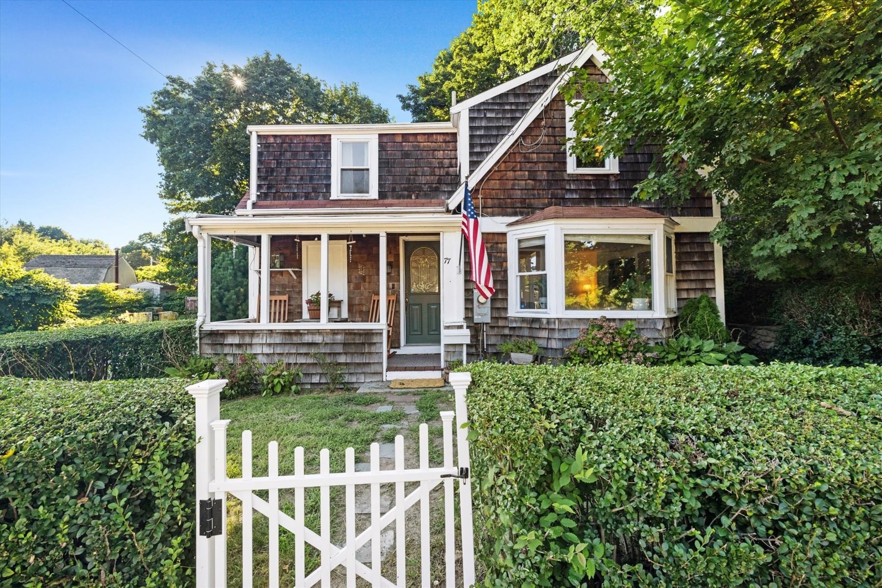 Shingle-sided house with a covered porch, bay window, American flag, and white garden gate.