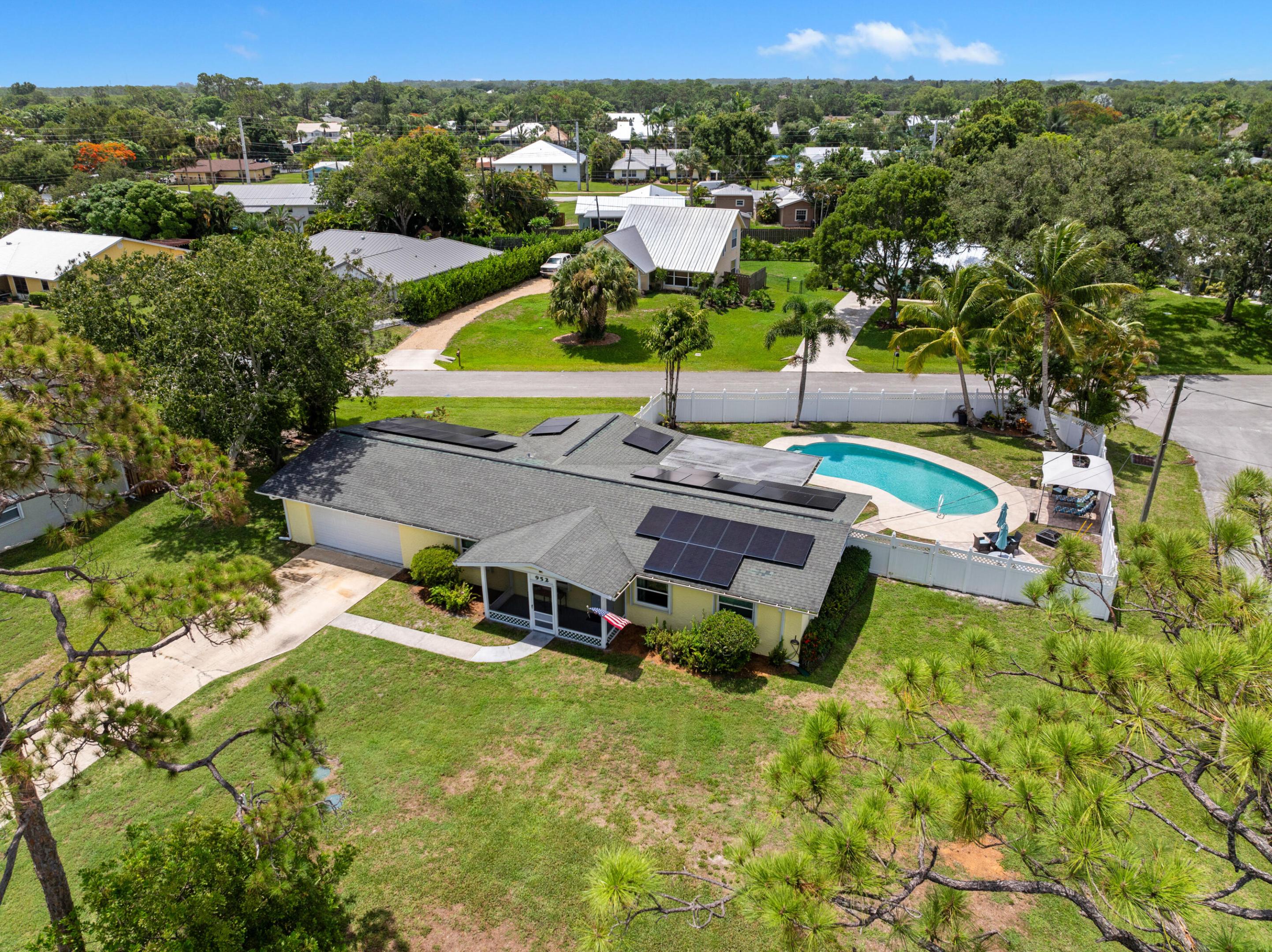 Aerial view of a single-story home with rooftop solar panels, a fenced pool, and a large lawn.
