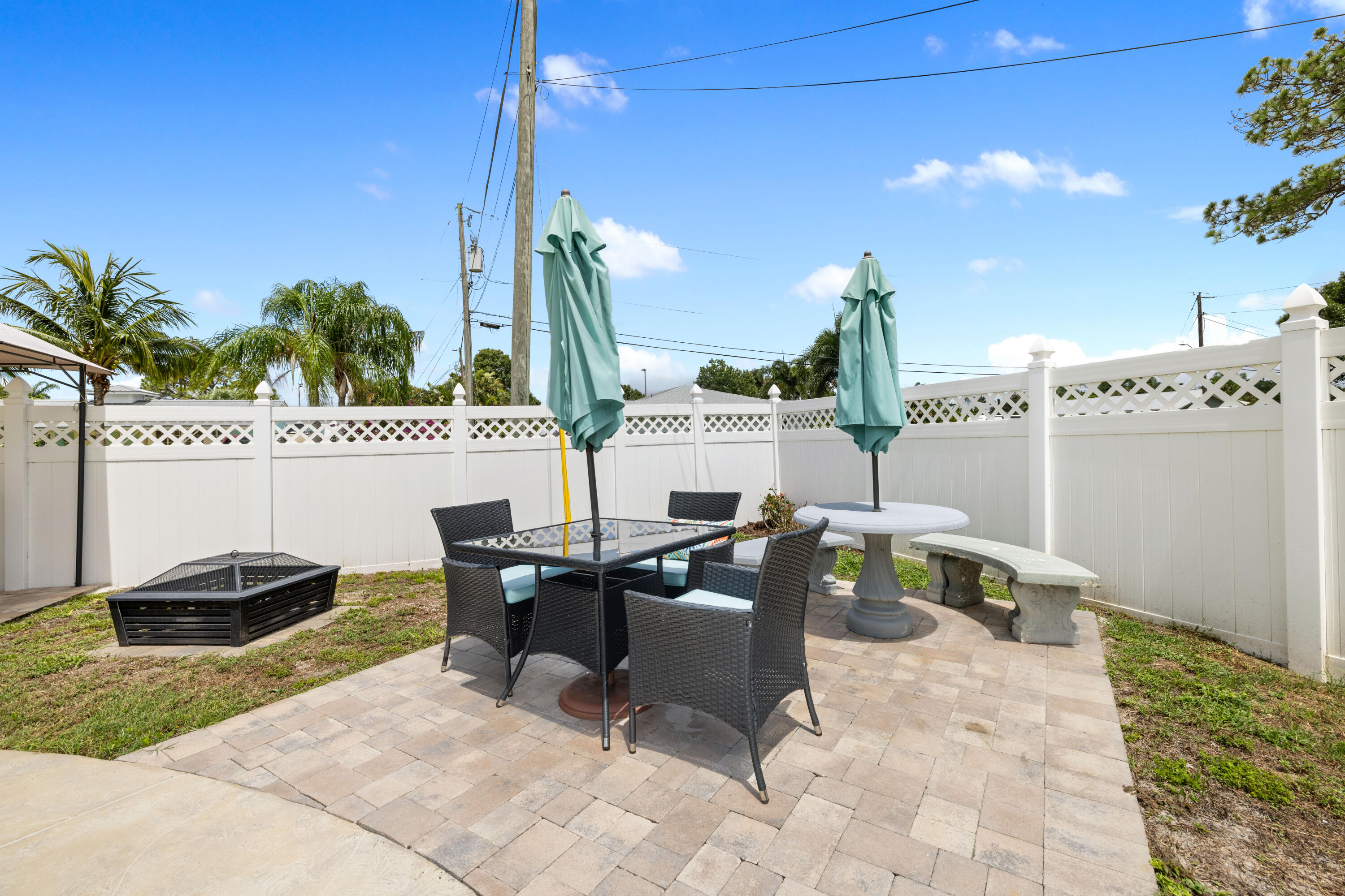 Backyard patio with wicker dining set, teal umbrellas, stone table and benches, and a white privacy fence.