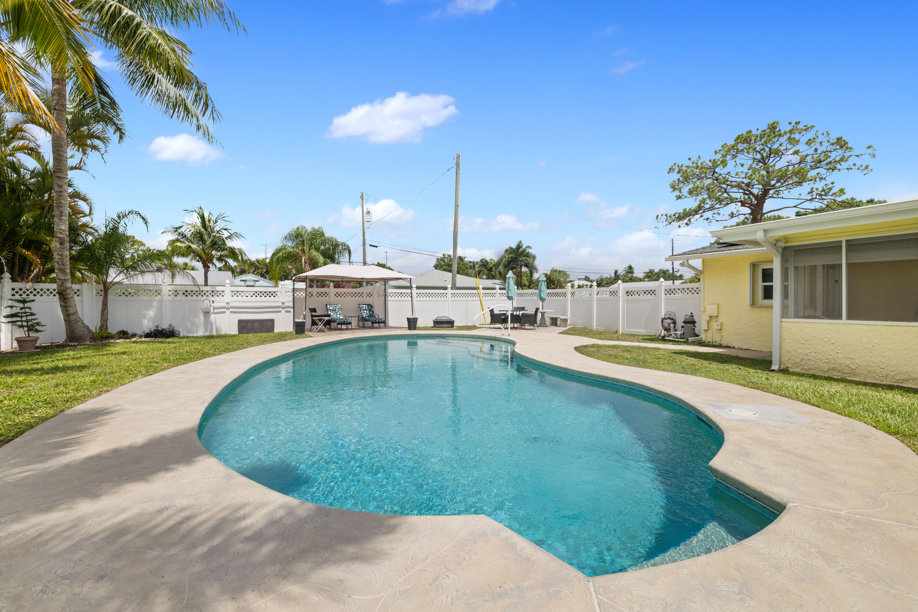 Curved backyard swimming pool beside a yellow house, with a fenced lawn and palm trees.