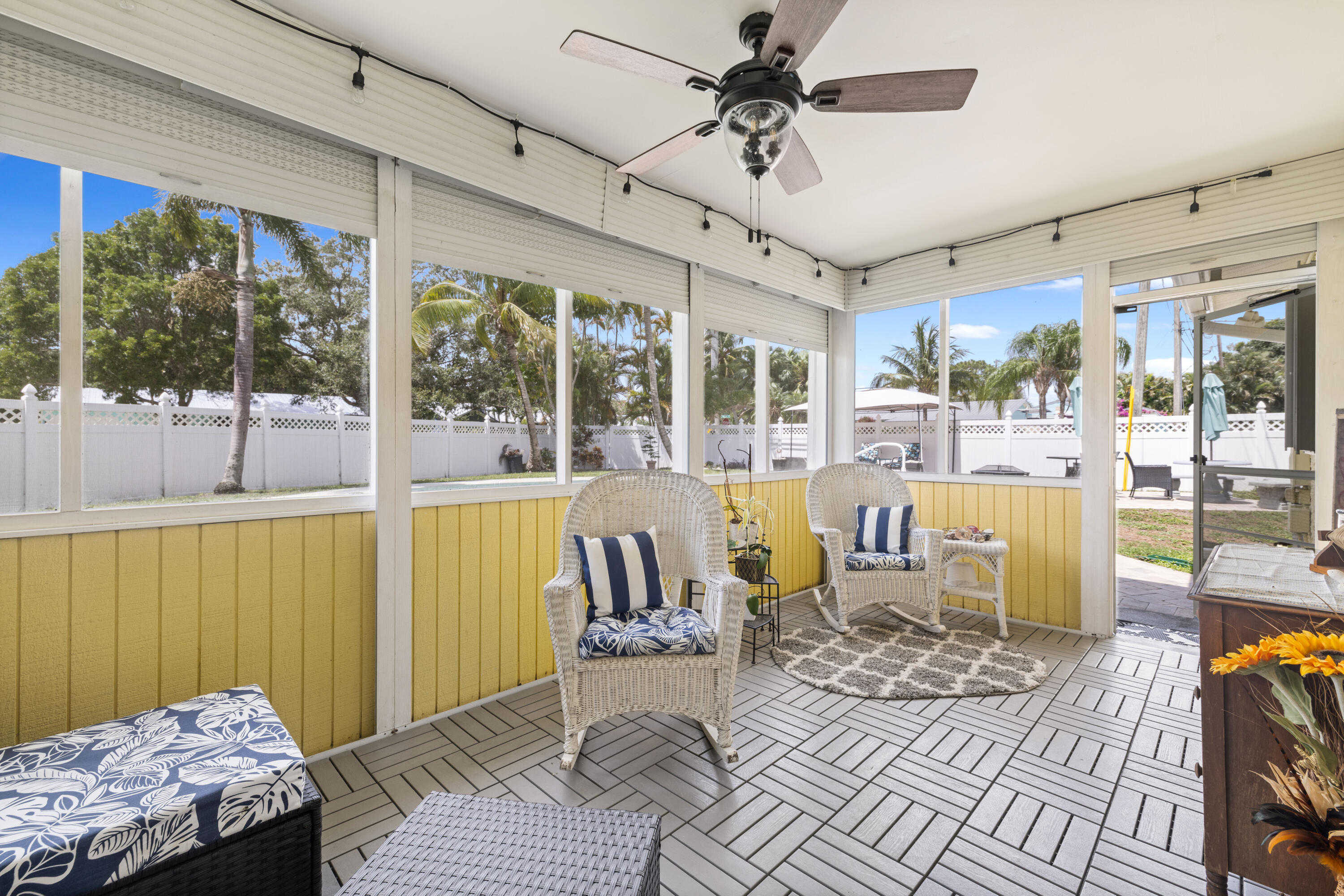 Enclosed sunroom with wicker chairs, yellow wall panels, and windows overlooking a tropical yard.