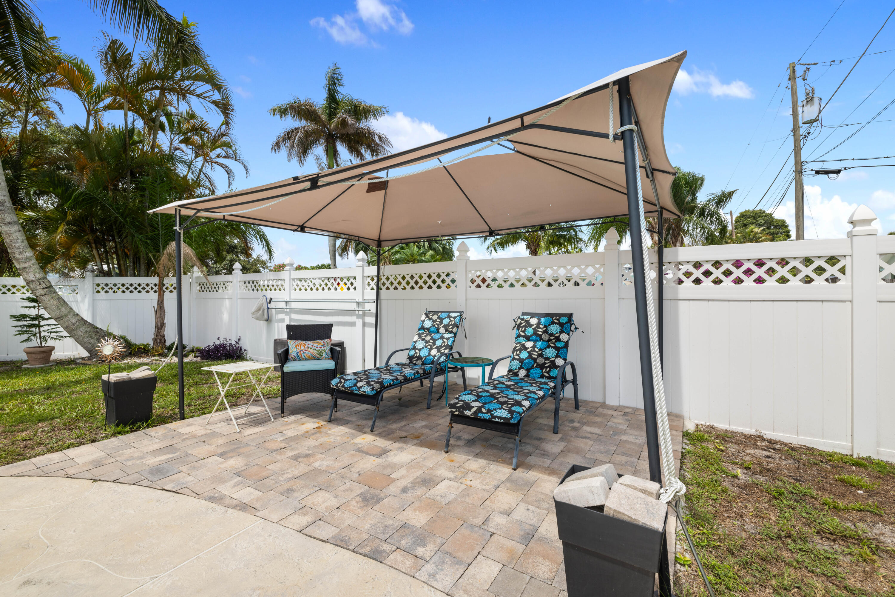Backyard patio with a large shade canopy, patterned lounge chairs, brick pavers, and a white fence.