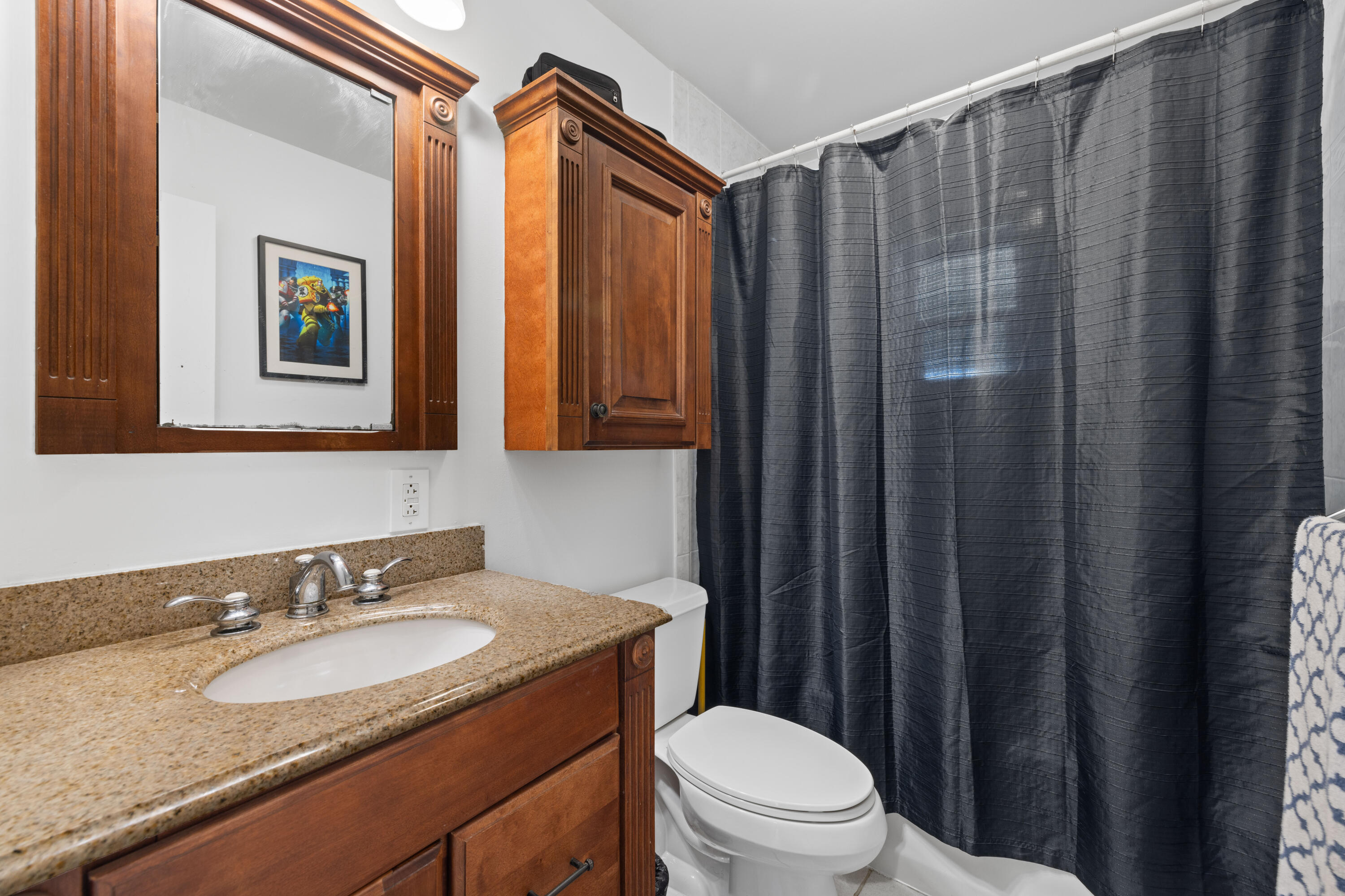 Bathroom with a granite vanity, dark wood cabinetry, white toilet, and charcoal shower curtain.