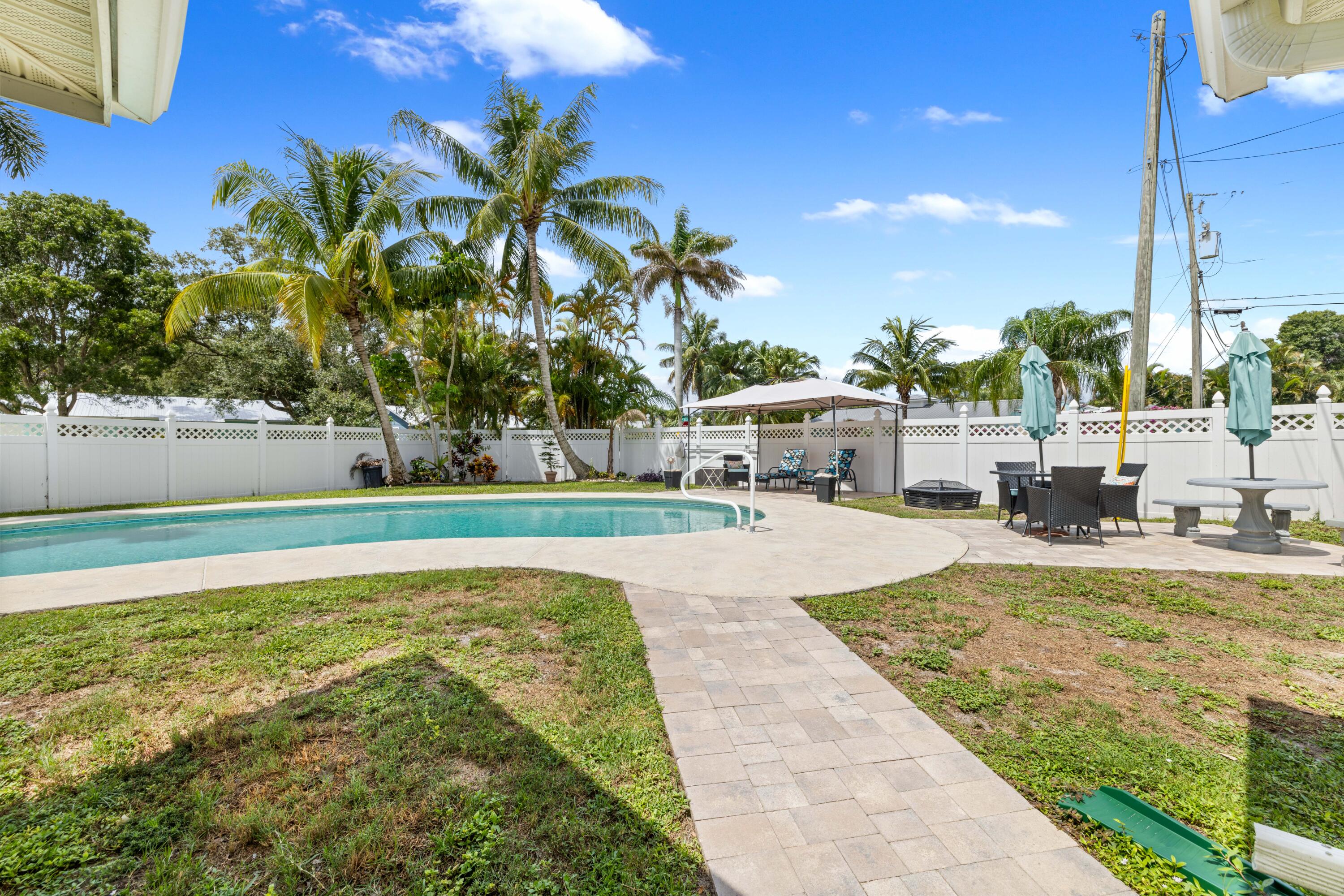 Backyard pool and paved patio with palm trees, outdoor seating, and a white privacy fence.