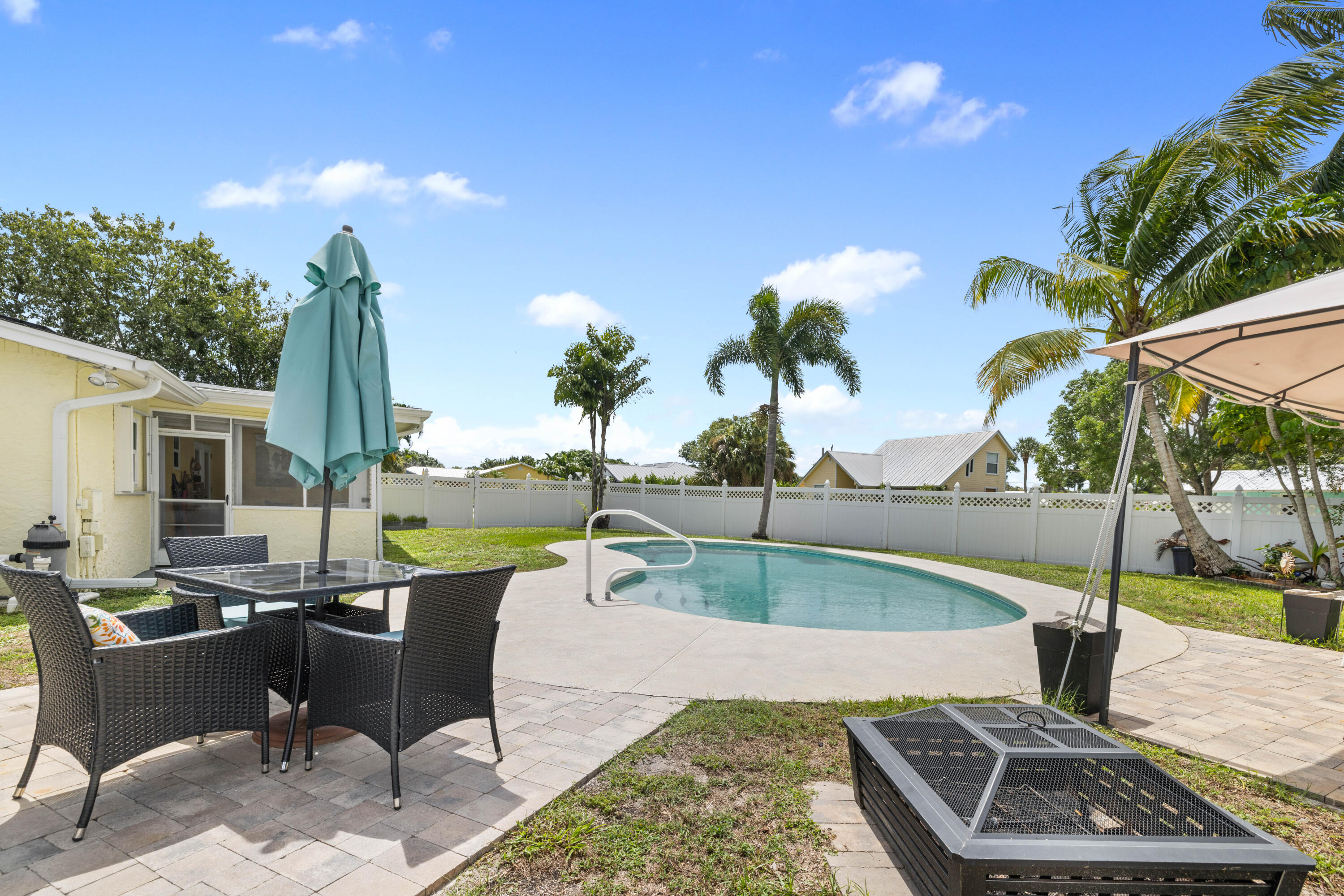 Backyard pool with a curved shape, paved patio, wicker dining set, and palm trees.