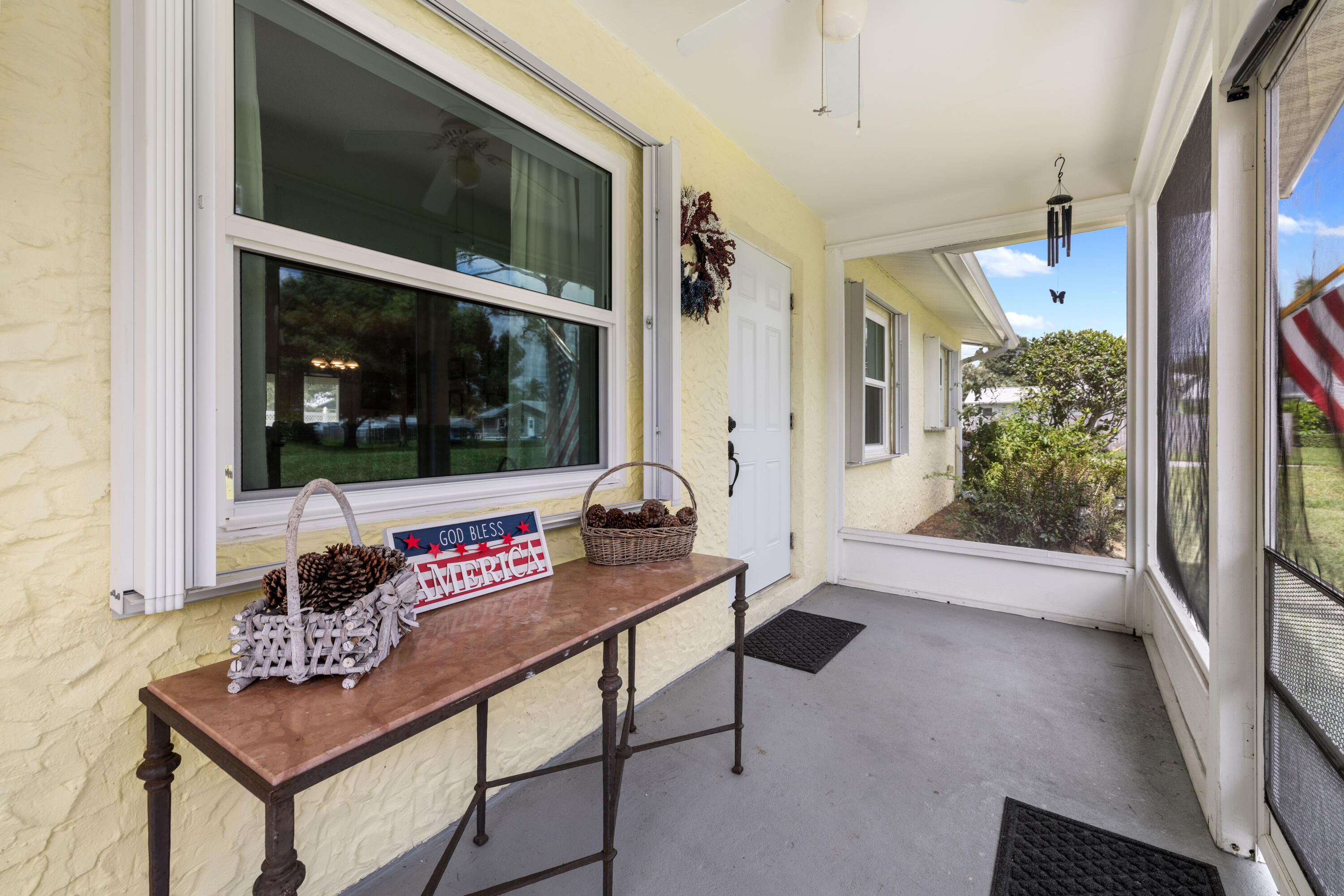 Screened front porch with a yellow stucco wall, white door, and table displaying a God Bless America sign.
