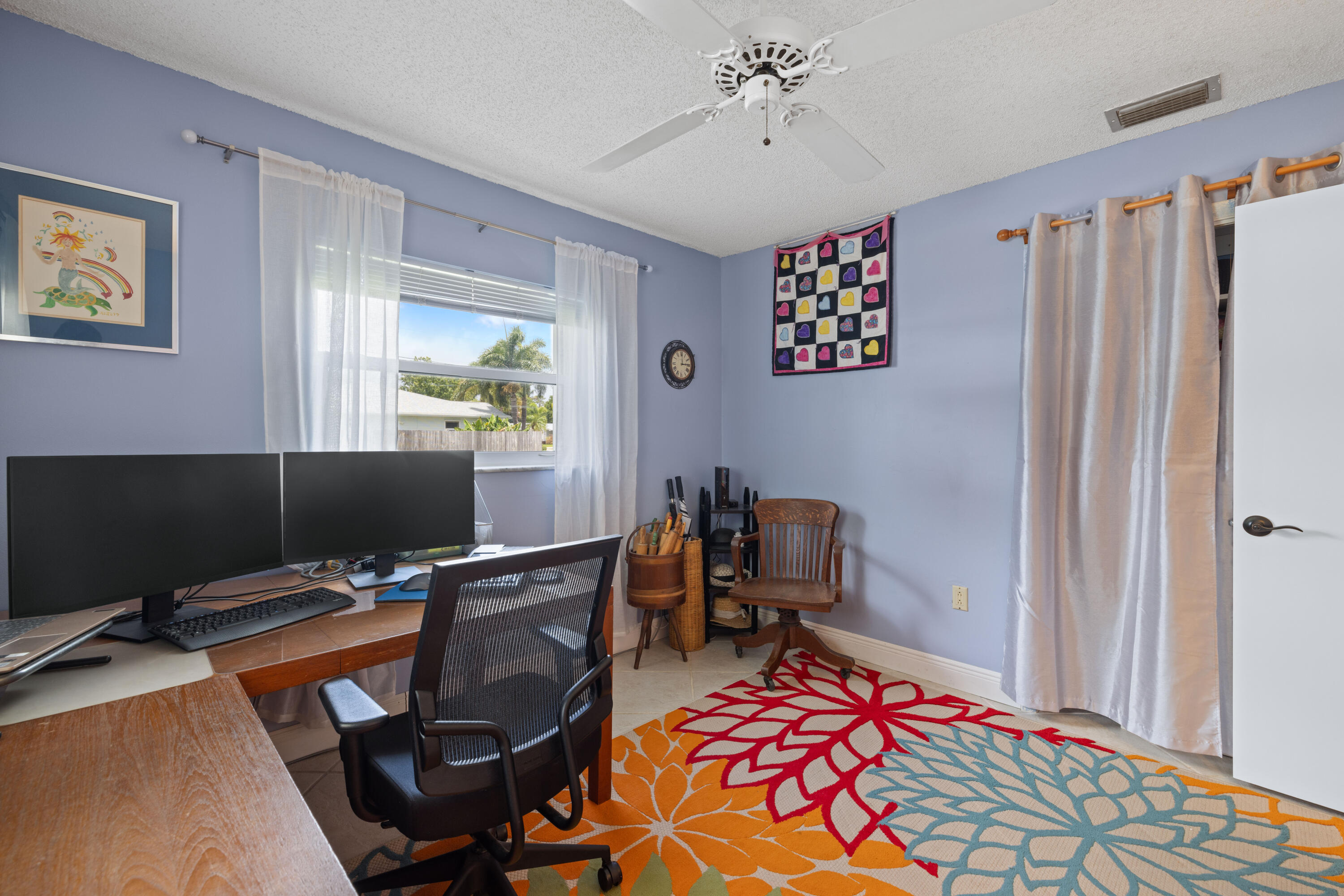 Home office with dual monitors, a wooden desk, lavender walls, and a colorful floral rug.