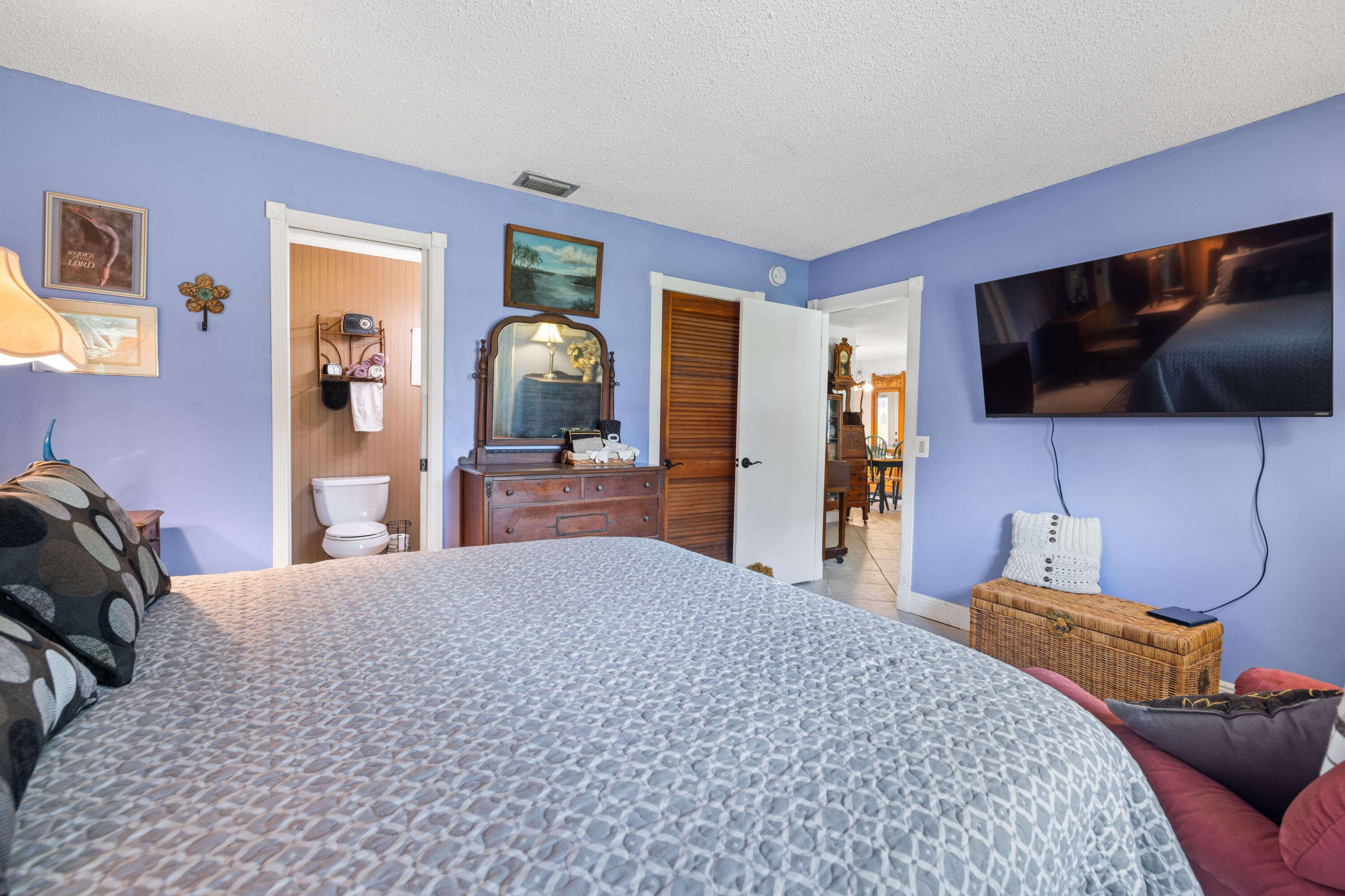 Bedroom with lavender walls, gray patterned bedding, a wooden dresser and mirror, and a wall-mounted television.