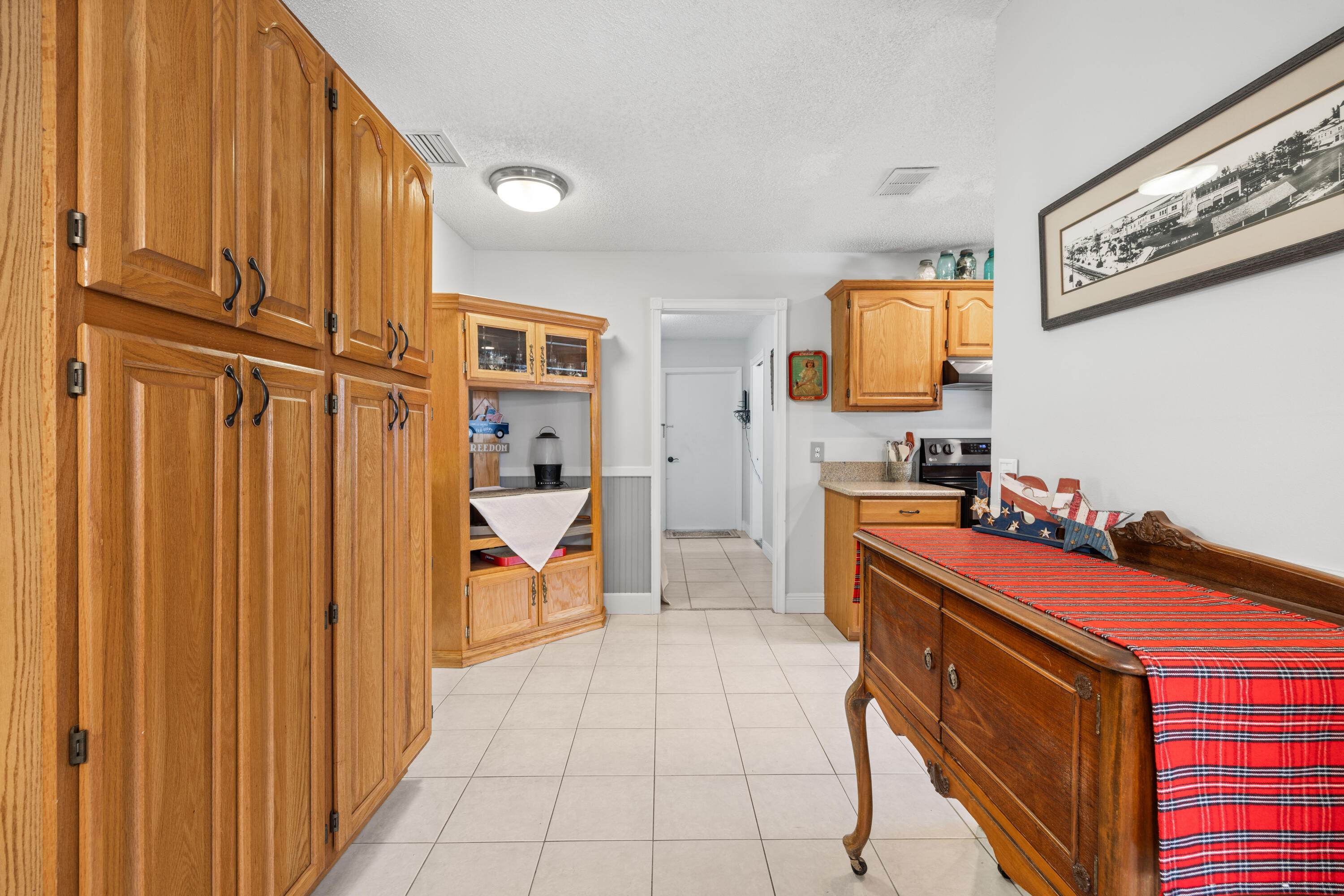 Kitchen with honey-colored wood cabinets, tiled floors, and a dark wood sideboard with a red plaid runner.