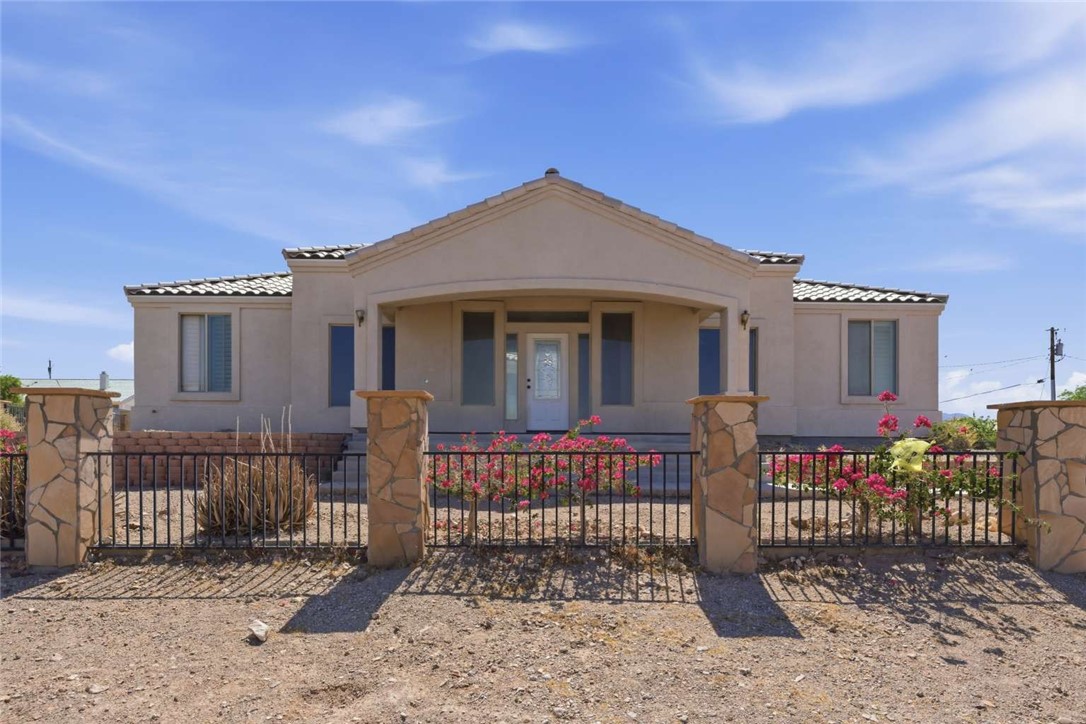Single-story stucco home behind a black metal fence with stone pillars and flowering shrubs.