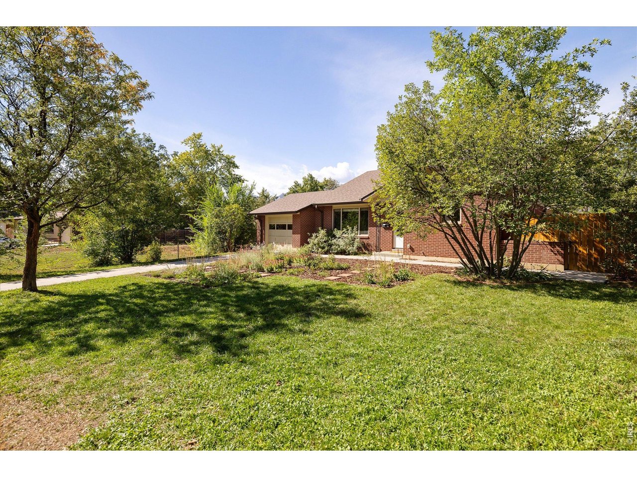 Red brick house with an attached garage, garden beds, mature trees, and a broad lawn.