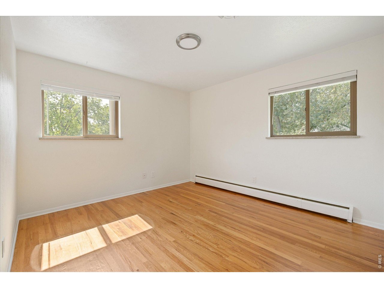 Empty room with hardwood floors, two windows, and a baseboard heater.