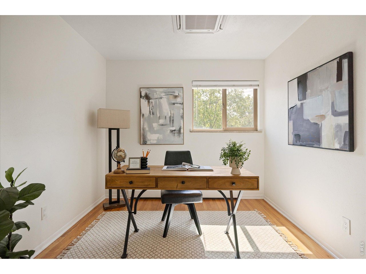 Staged home office with a wooden desk, black chair, patterned rug, and window overlooking trees.