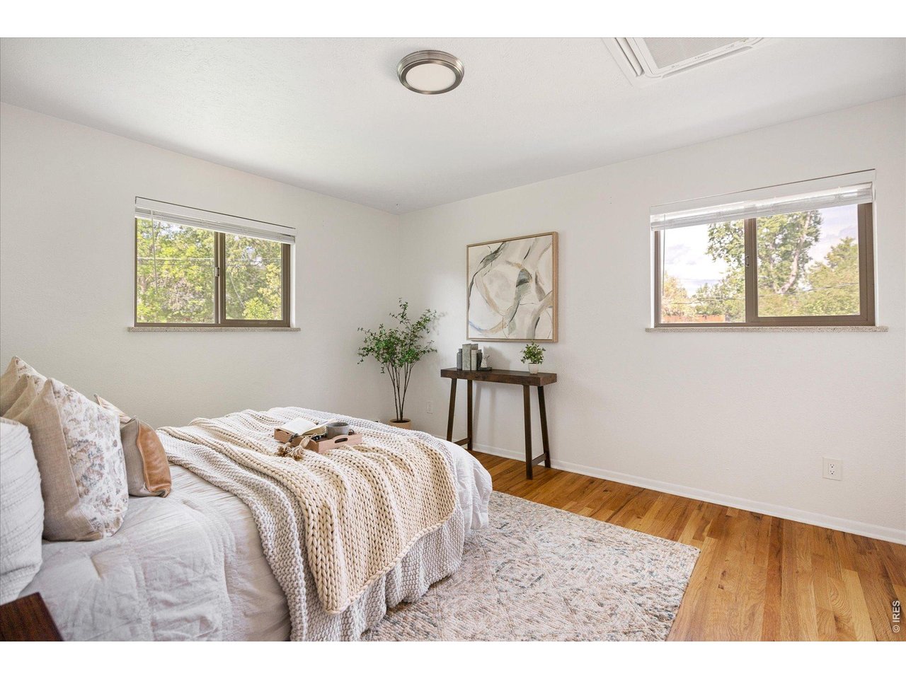 Staged bedroom with a white bed, knit blanket, two sunny windows, and hardwood floors.