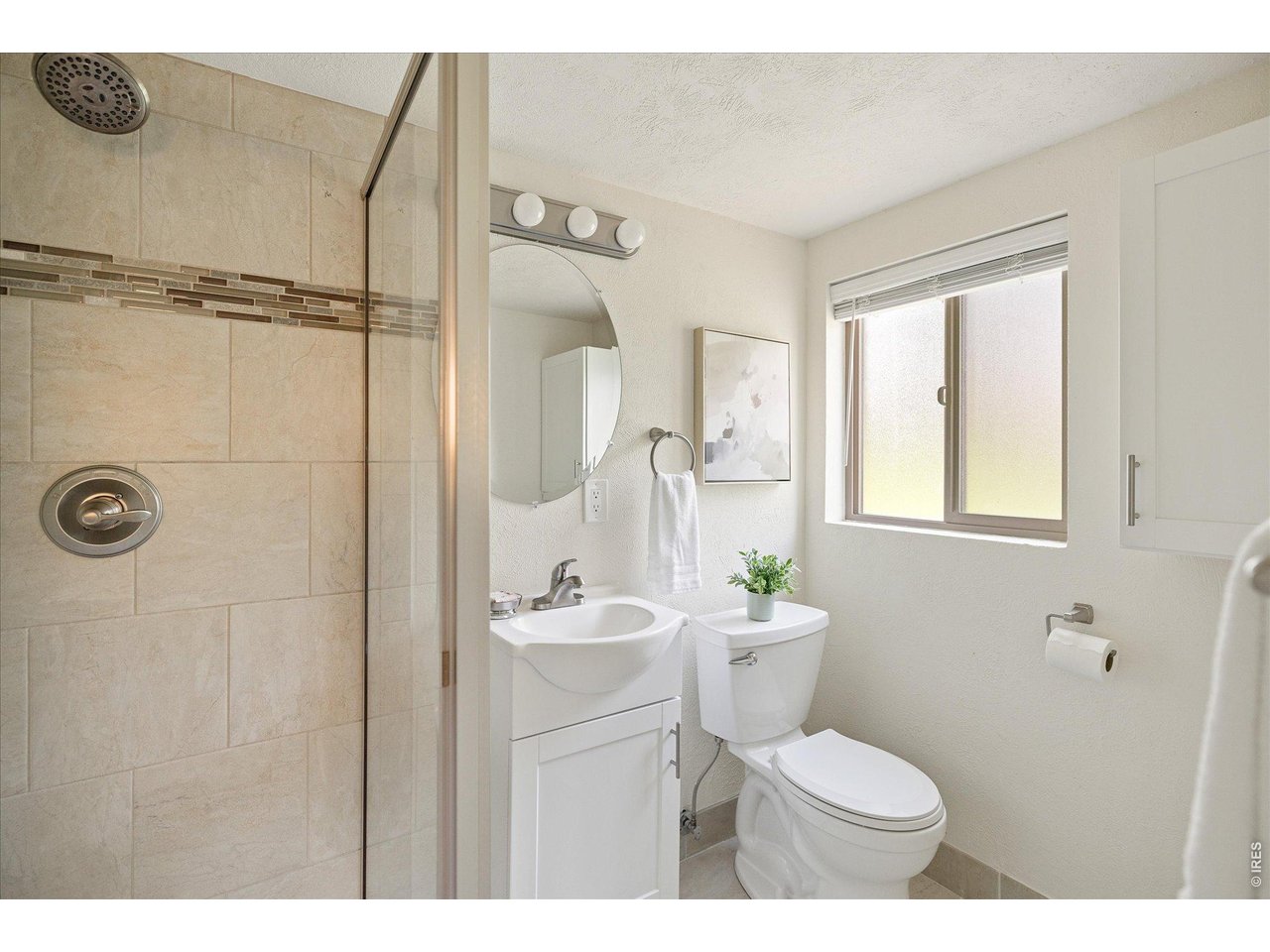 Bathroom with a beige tiled shower, white vanity, rounded mirror, and window.