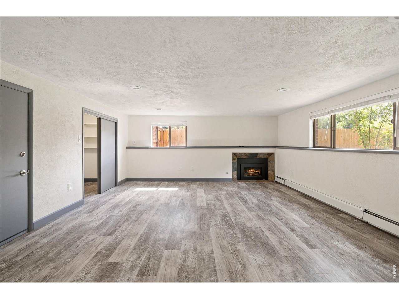 Empty room with gray plank flooring, a small fireplace, and windows facing a fenced yard.