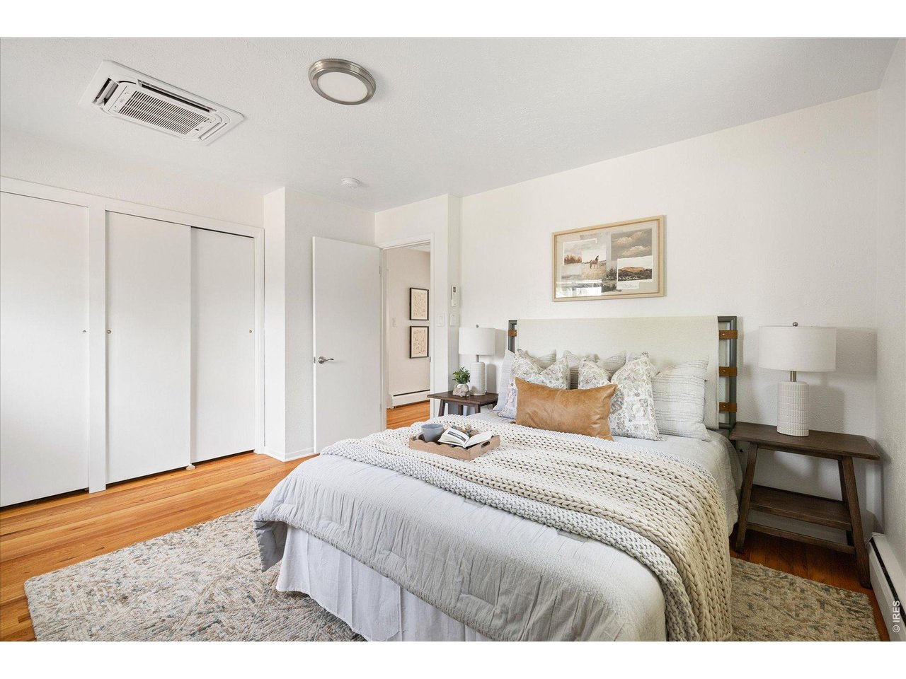 Staged bedroom with a light upholstered bed, white sliding closet doors, and warm hardwood floors.