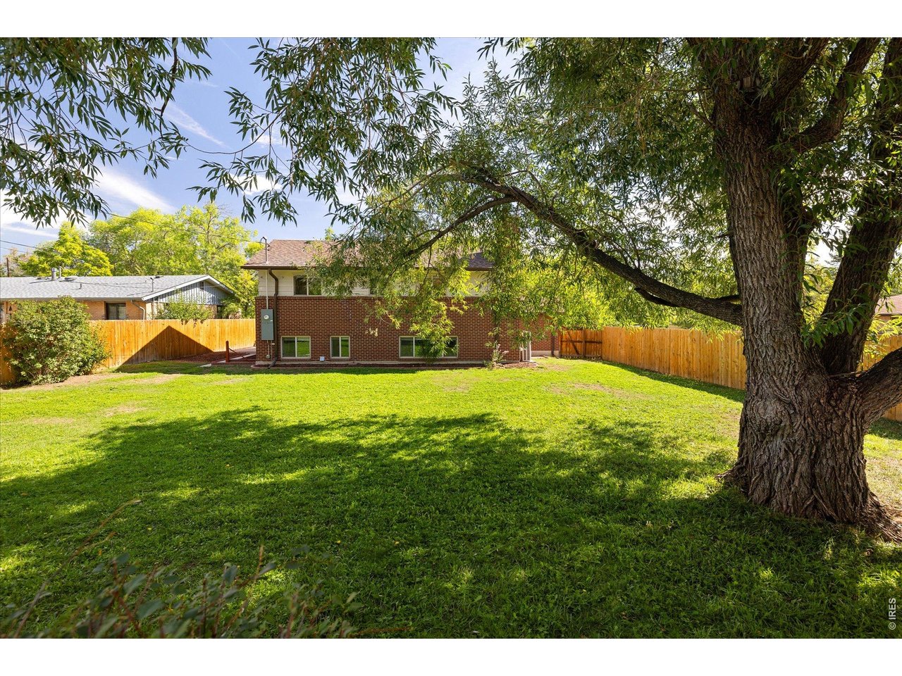 Rear of a brick home behind a fenced lawn, shaded by a large mature tree.