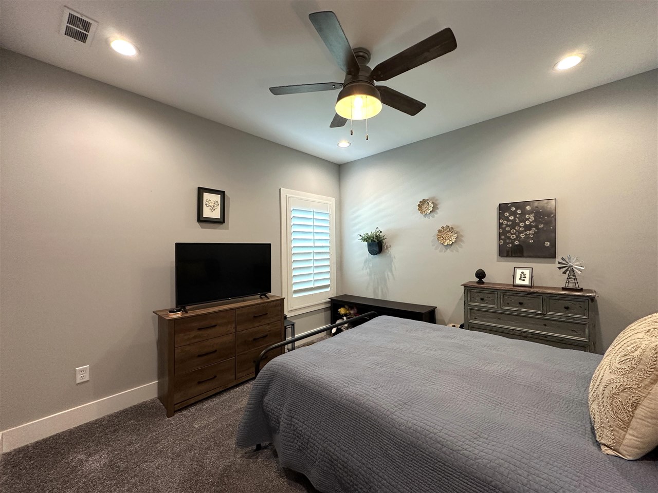 Furnished bedroom with a gray bedspread, ceiling fan, shuttered window, and wooden dressers.