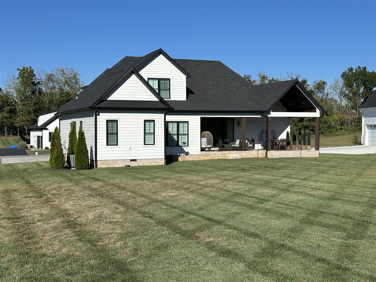 White gabled house with dark trim, covered furnished patio, brick foundation, and broad lawn.