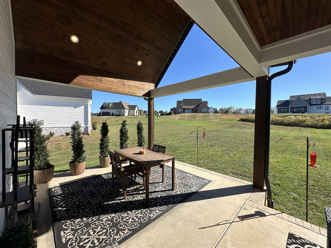 Covered patio with a wood ceiling, outdoor dining set, potted evergreens, and a view of the grassy yard.