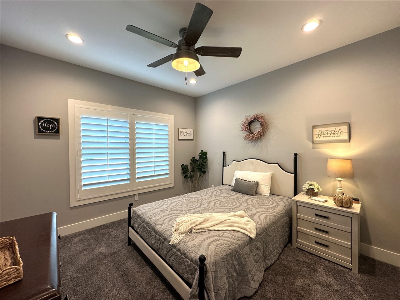 Furnished bedroom with a black metal bed, patterned gray bedding, white shutters, and a ceiling fan.