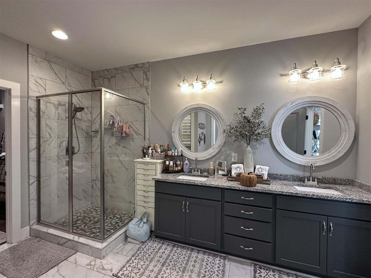 Bathroom with a marble-look walk-in shower and dark double vanity with round mirrors.