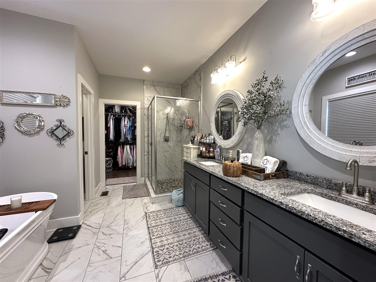 Primary bathroom with a long double vanity, round mirrors, marble-look shower, and freestanding tub.