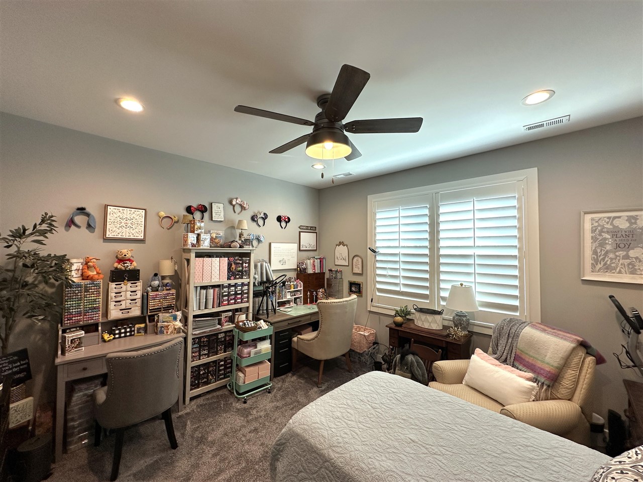 Bedroom with a bed, craft-supply shelves, a sewing desk, and plantation shutters.