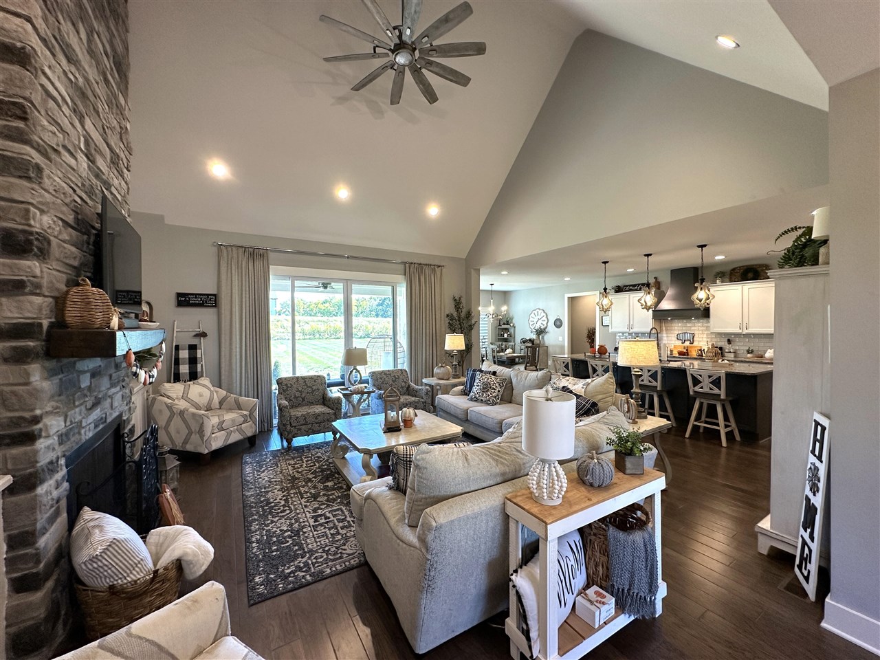 Staged living room with a vaulted ceiling, stone fireplace, and open kitchen.
