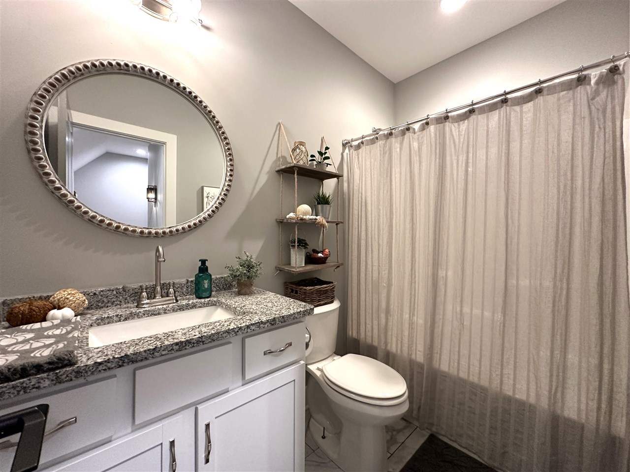 Bathroom with a granite vanity, oval mirror, toilet, and light fabric shower curtain.