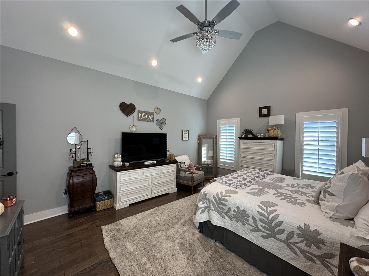 Bedroom with a vaulted ceiling, crystal ceiling fan, white dressers, and plantation shutters.