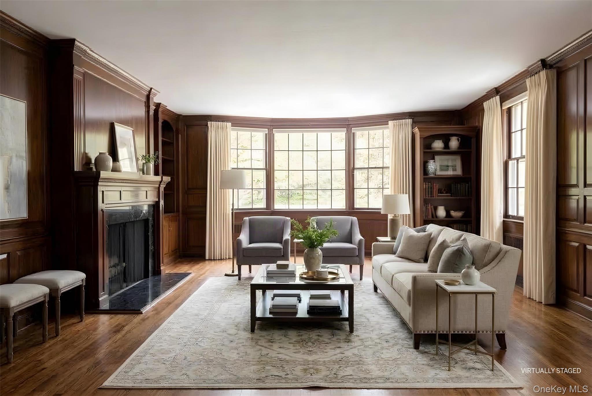 Living room with dark wood paneling, a fireplace, and large bay windows, virtually staged with neutral furnishings.