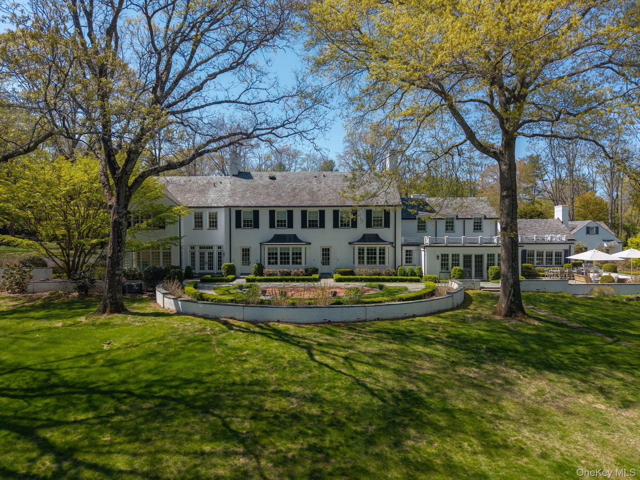 Large white colonial home framed by mature trees, with a circular garden and patio umbrellas.