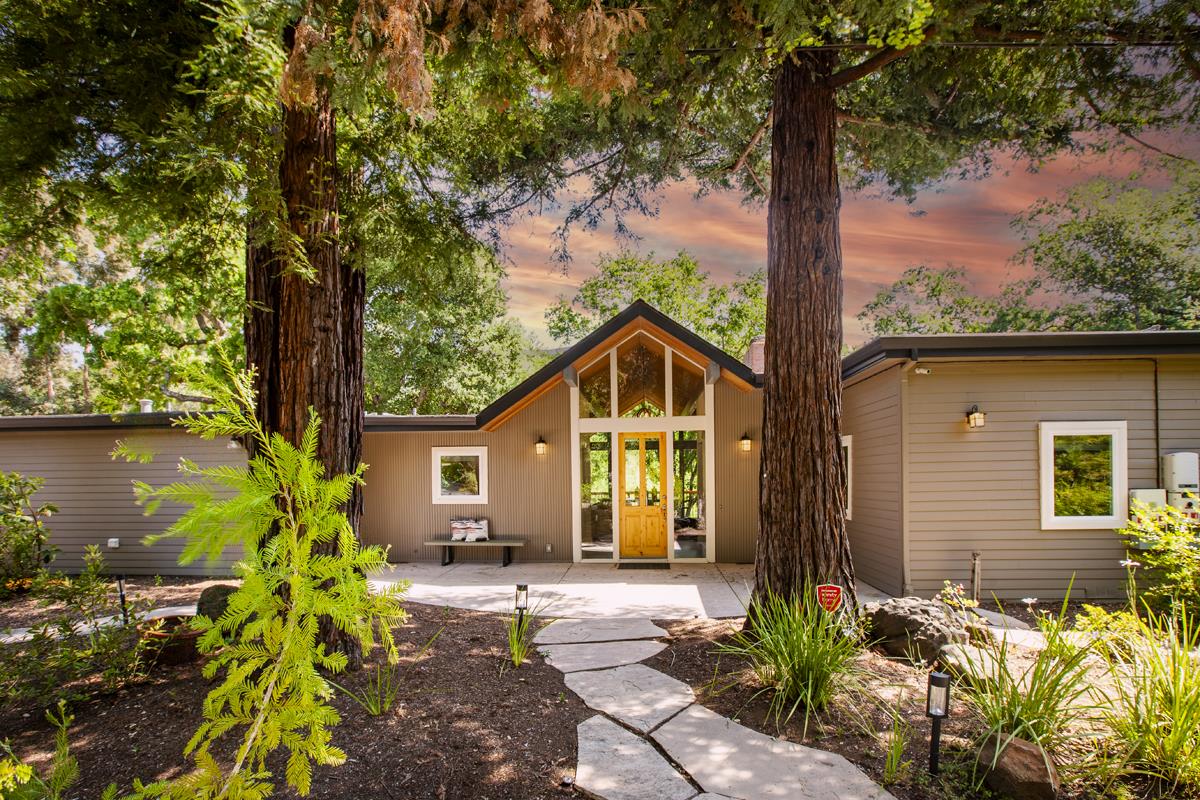 Home with a peaked glass entry and yellow door, approached by a curving stone walkway beneath tall trees.