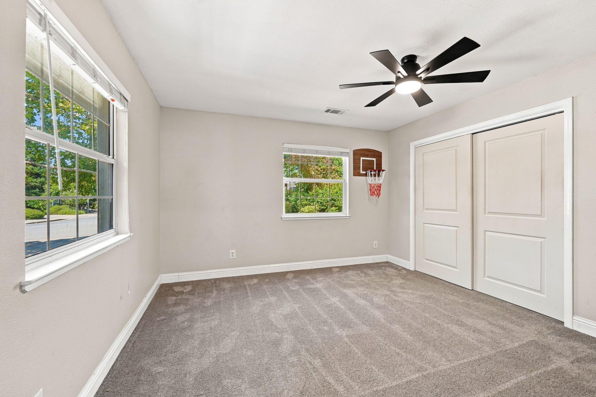Carpeted room with large windows, a ceiling fan, closet doors, and a basketball hoop.