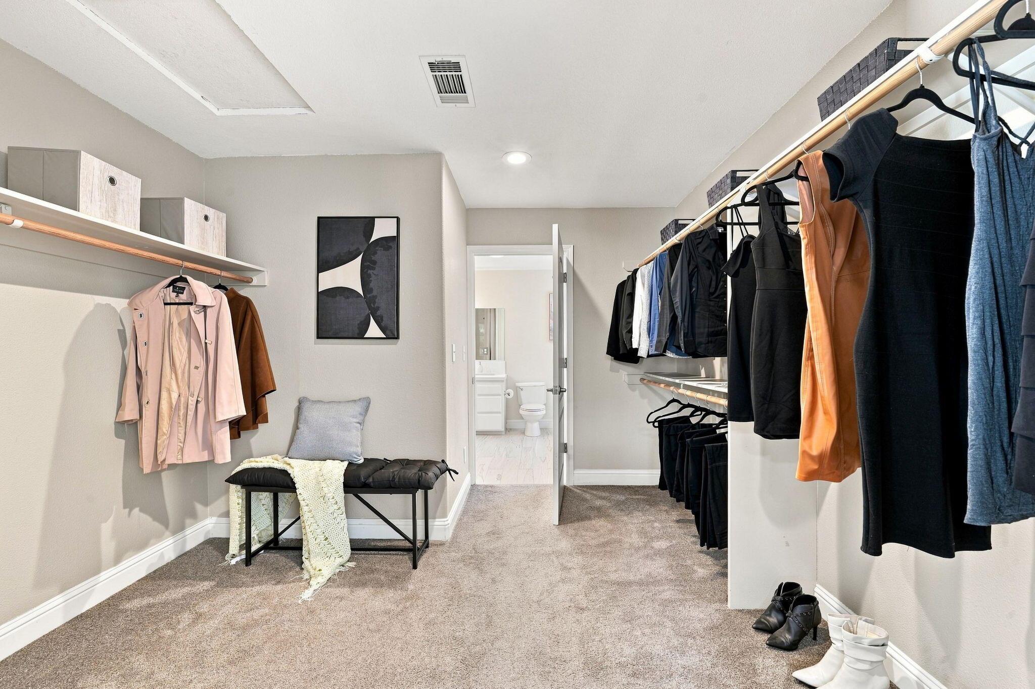 Walk-in closet with clothing on open racks, a cushioned bench, and a bathroom doorway.