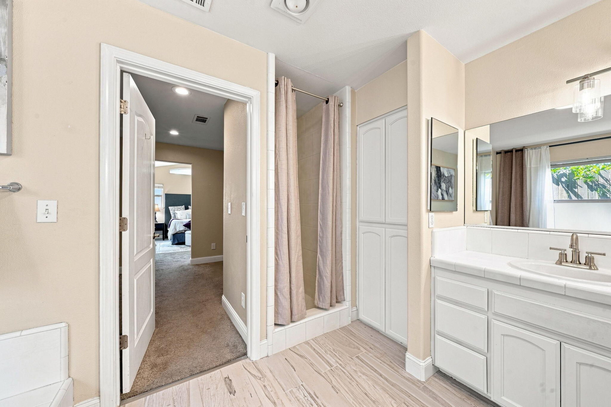 Bathroom with a white vanity, tiled shower, and built-in cabinets beside an open bedroom doorway.