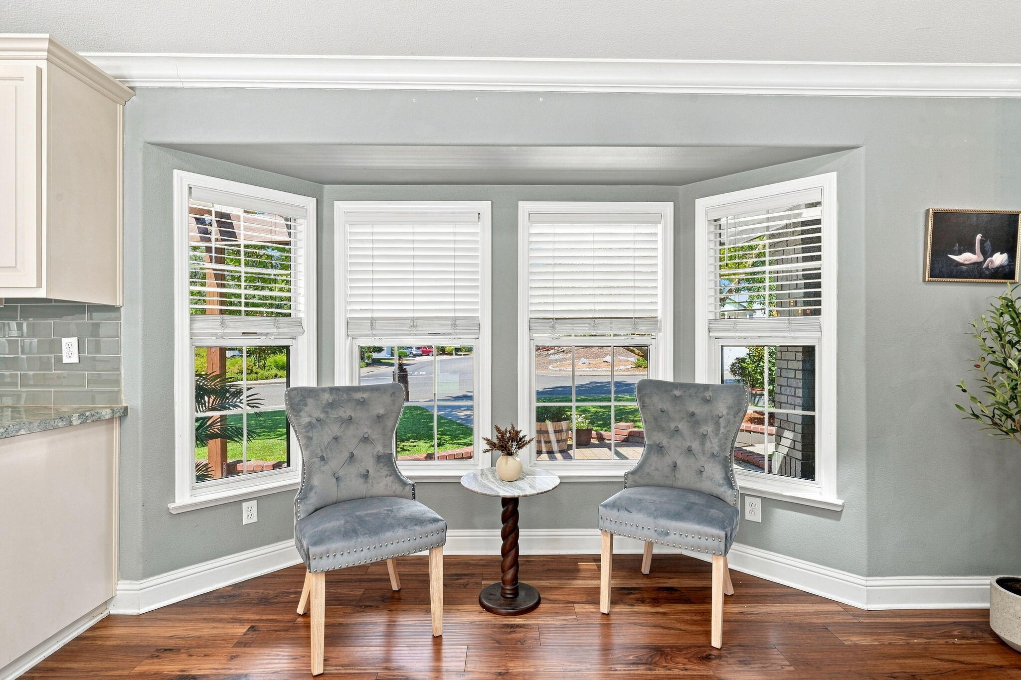Bright sitting area with two gray chairs and a small table in front of bay windows.