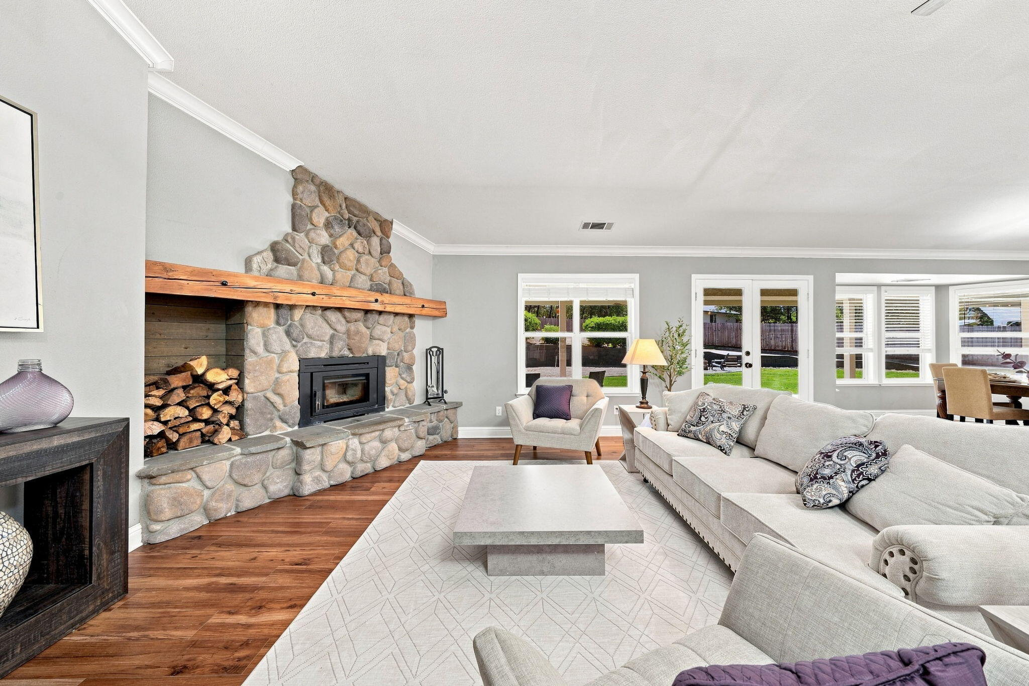 Living room with a stone fireplace, wood mantel, pale sectional, and glass doors opening to the backyard.
